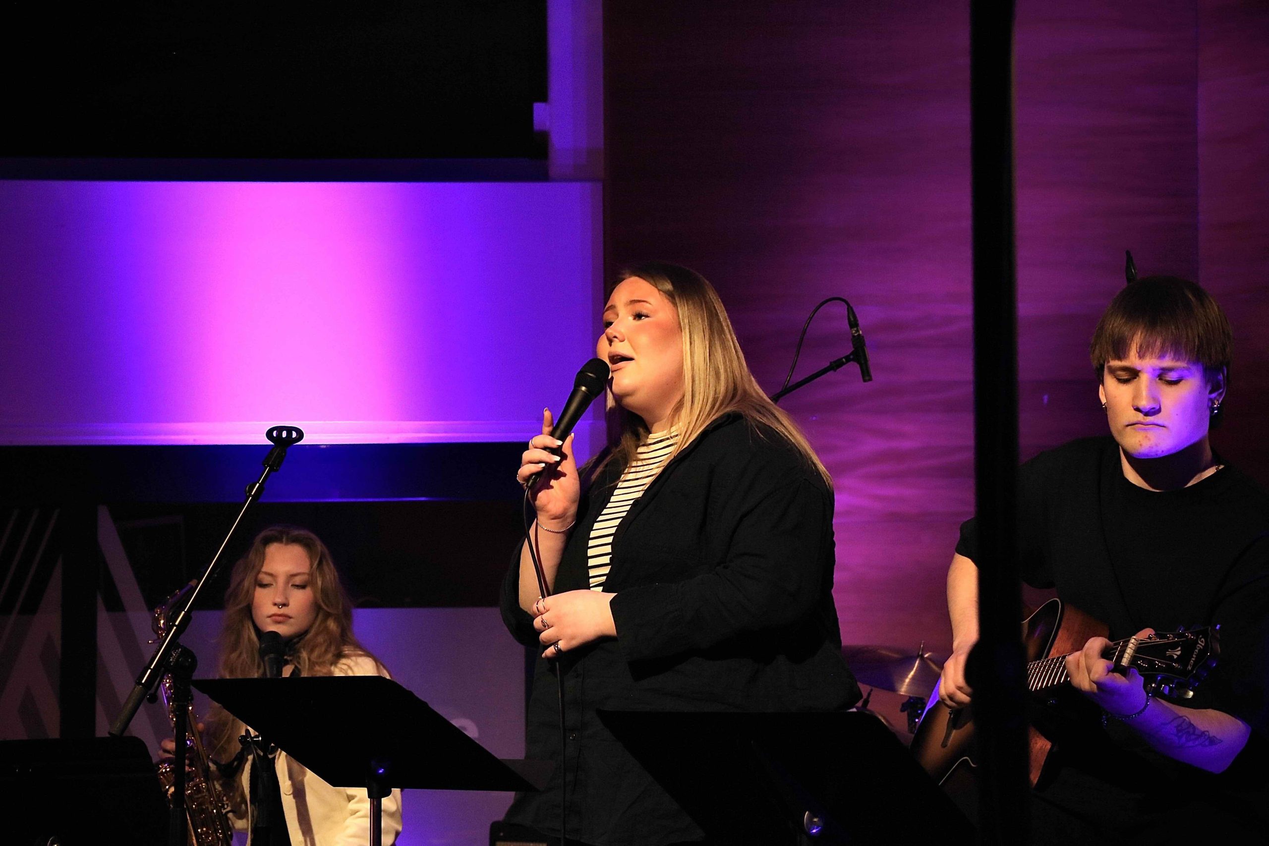 En ung sångerska längst fram i scen, en ung saxofonist som sitter i bakgrunden och håller saxofonen i ena handen.