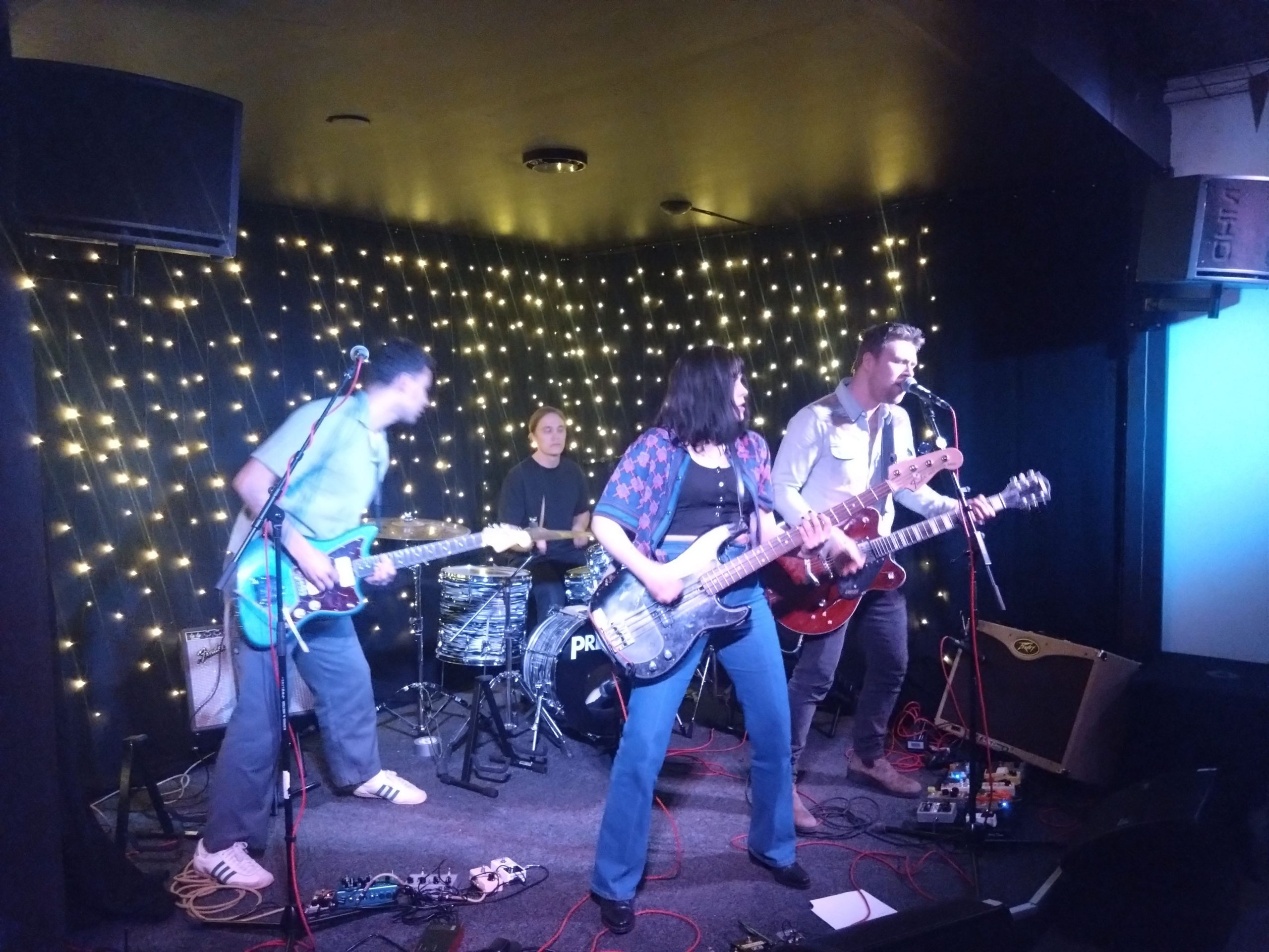 Asian Cowboys basist står längst fram i mitten på Kajmans scen, till vänster om henne står gitarristen och till höger om henne står frontpersonen som spelar på sin brunröda elgitarr och sjunger i mikrofonen. Längst bak sitter trummisen bakom trumsetet.