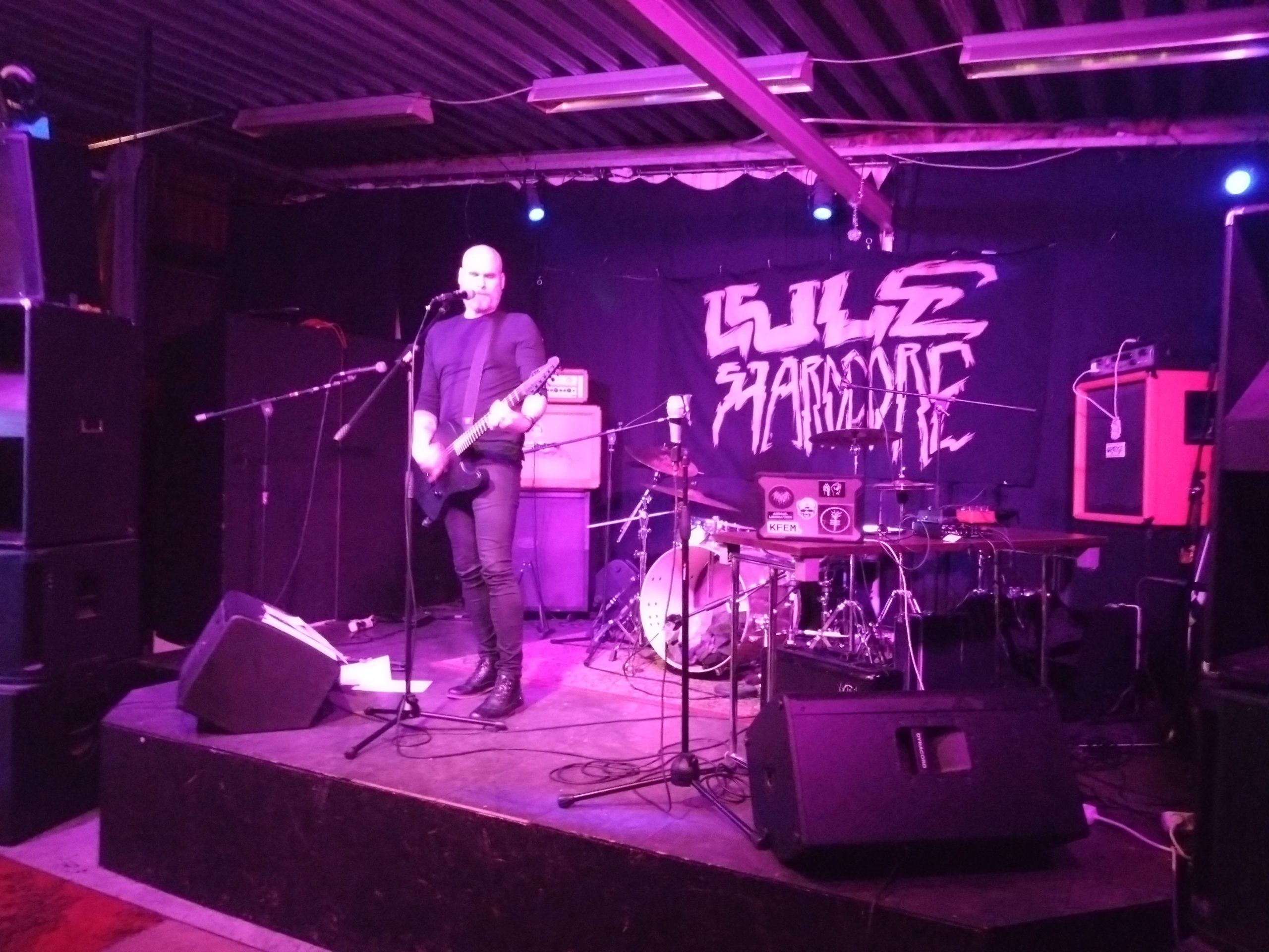 Artisten Vita Bruset står ensam på scenen med sin svarta elgitarr och en mikrofon framför sig. Han har svarta kläder, rakat huvud och står i rosa scenbelysning. Det ser ganska ödsligt och tomt ut framför scenen.