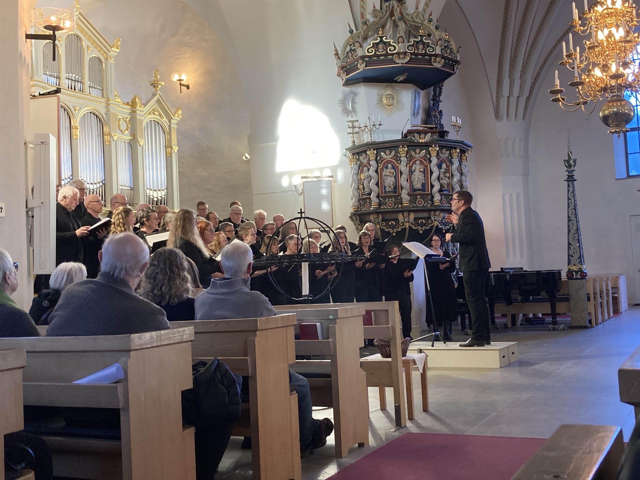 Kören och dirigenten som är mitt uppe i att sjunga stycket. Bilden är tagen från en kyrkbänk. Öjebyns kyrka har vitmålade väggar, välvda tak och ljuskronor i mässing. Kyrkan har många dekorativa inslag med olika religiösa utsmyckningar.