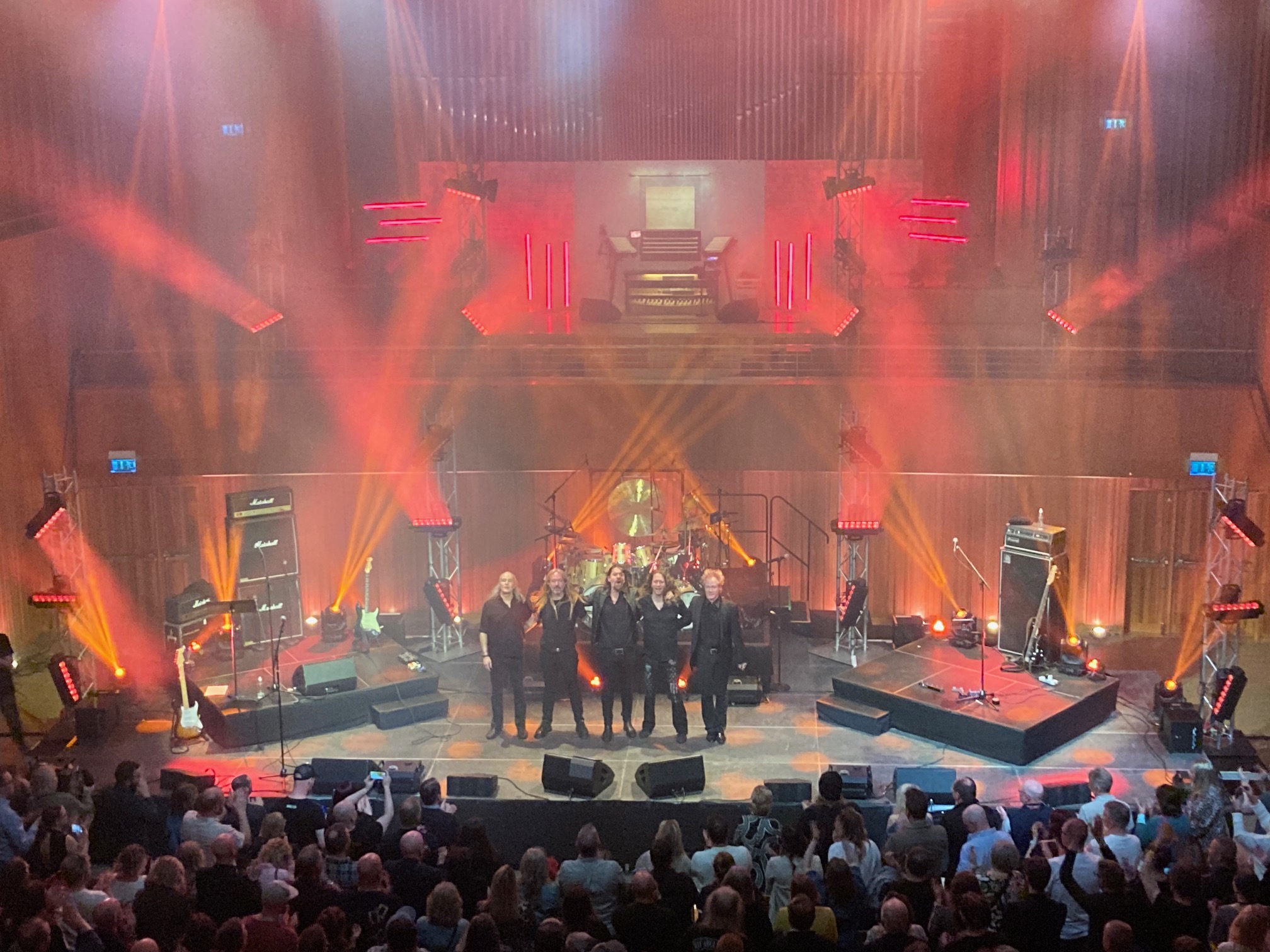 De medverkande musikerna står uppradade på scenen och tar emot publikens applåder. Framför scenen står publiken och ger stående ovationer.