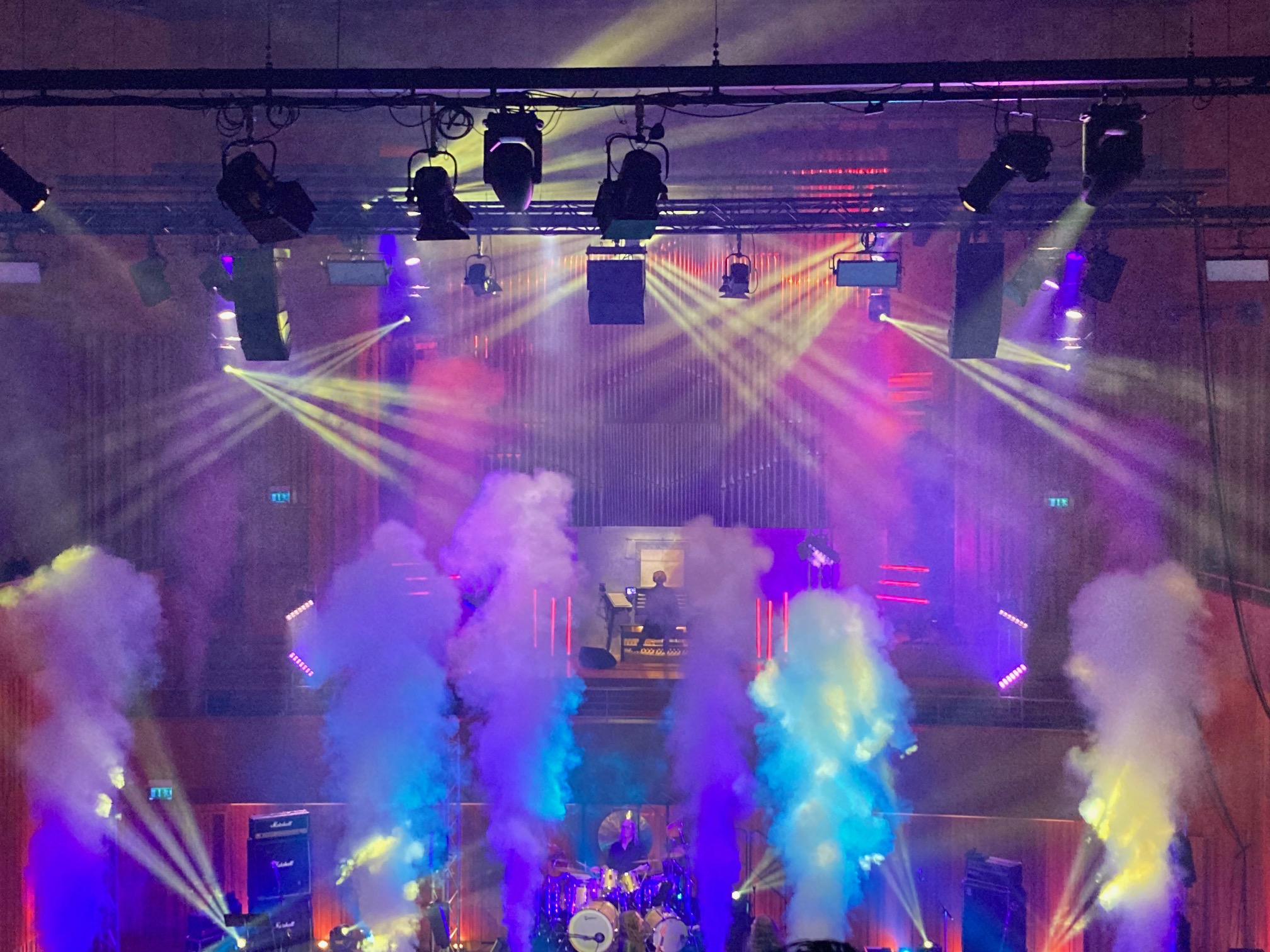 Musikerna på scenen. Rökmaskiner, pyroteknik och effektfullt ljus i blått, rosa och lila. Uppe på andra våningen sitter organisten vid orgeln.