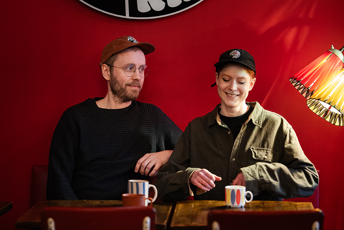 Norrativets projektledare Josef Eriksson till vänster och Linn Kårelind till vänster. De sitter vid ett bord med varsin randig kaffekopp framför sig. Josef har brun keps, glasögon och skägg och han tittar snett mot Linn. Linn har svart keps, piercing i näsan och hon tittar ner i bordet skrattandes.