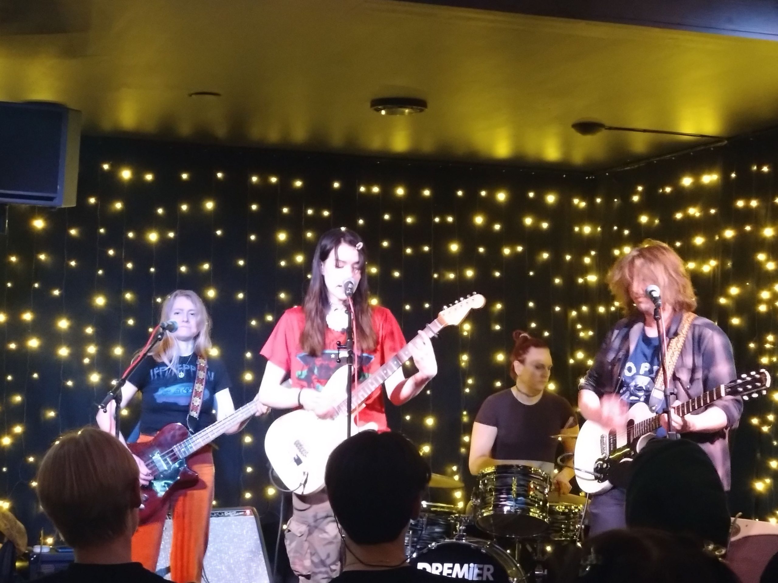 Frontfiguren Linn står i mitten på Kajmans scen med sin vita elgitarr och sjunger i mikrofonen. Bakom henne står den kvinnliga basisten, till höger om Linn står den manliga gitarristen och längst bak sitter trummisen bakom trumsetet. Alla ser fokuserade ut.