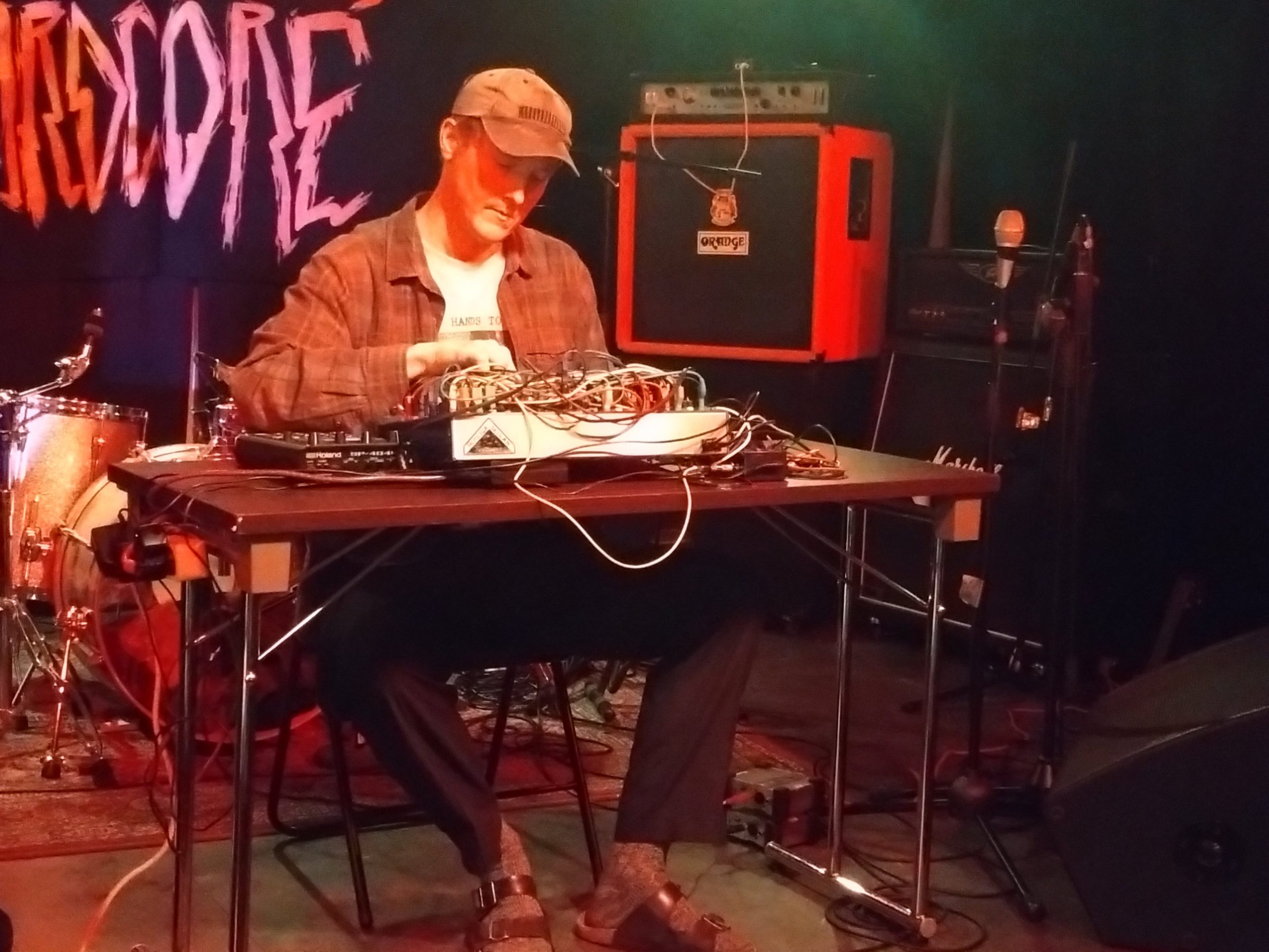 Artisten Märg sitter på scenen iklädd en brun, rutig skjorta, brun keps och svarta tofflor. På bordet framför sig har han ett mixerbord och synth och annan elektronik som han använder för sin musik. Han skruvar på mixerbordet och ser koncentrerad ut.