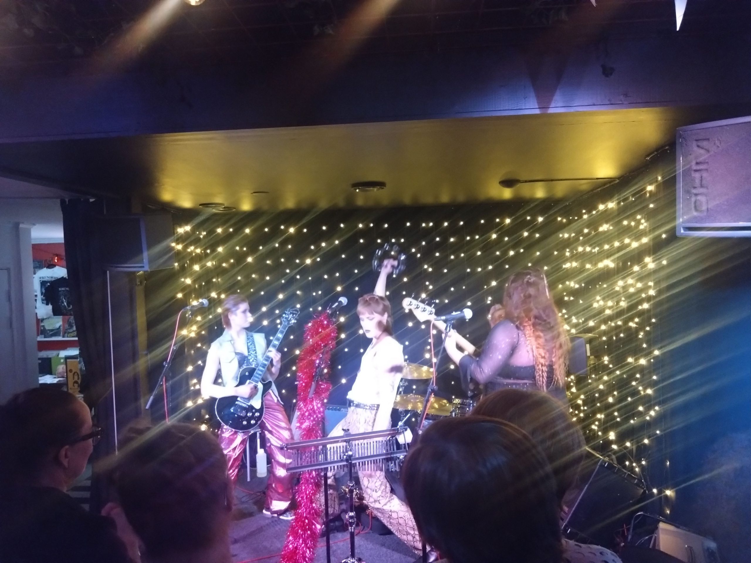 Rosa Blend har klätt det mittersta mikrofonstativet i rosa, glittrigt, lurvigt material. Frontfiguren står i mitten och håller upp en tamburin, till vänster om henne står gitarristen, som har en svart elgitarr, ljus jeansväst och rosa, blanka byxor i ett plastigt material, längst ut till höger står basisten vänd mot trummisen, som skymtas i bakgrunden.