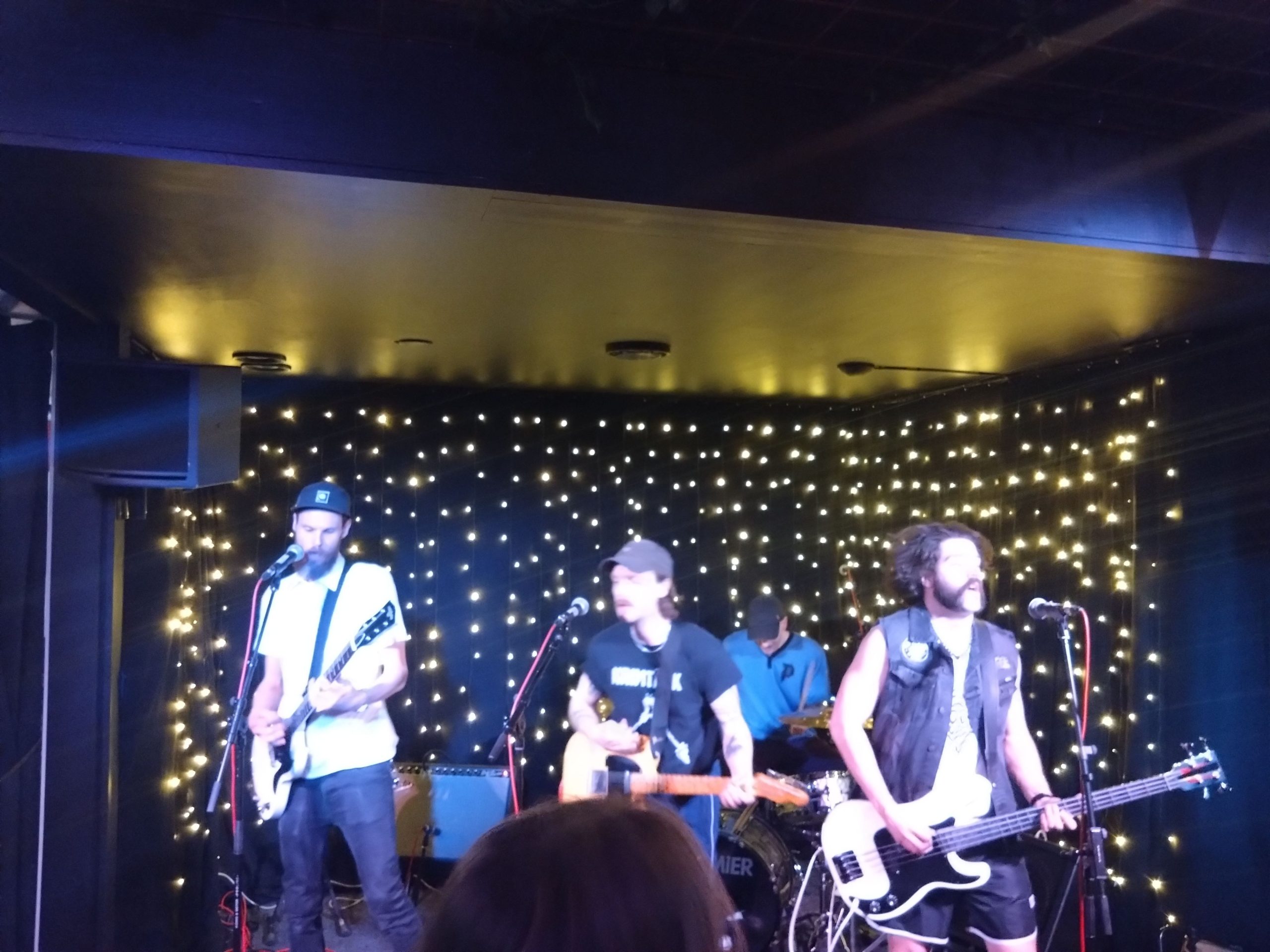 En suddig bild där Sekundernas frontfigur står längst fram med sin beiga elgitarr, han har keps och mustasch. Till vänster om honom står den andra gitarristen som också har keps. Längst ut till höger står basisten som har en vit och svart bas och längst bak skymtar man trummisen, som också har keps.