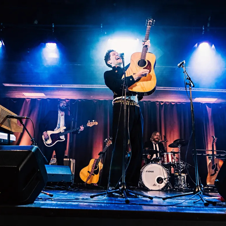 Live på en scen står Månskensbonden med en akustisk gitarr riktad uppåt
