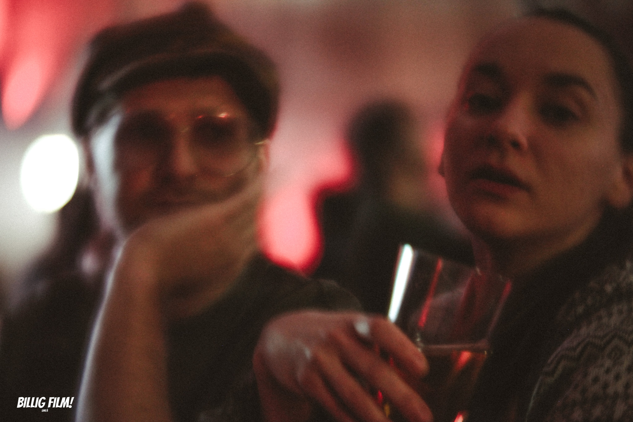 Två besökare på en blurrig mingelbild. Personen i förgrunden, en kvinna, håller ett göas och ser in i kameran. Mannen till vänster om henne har keps och glasögon och ser rakt in i kameran.