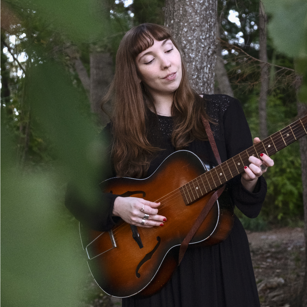 Mörkhårig kvinna med lugg står vid ett träd och spelar gitarr