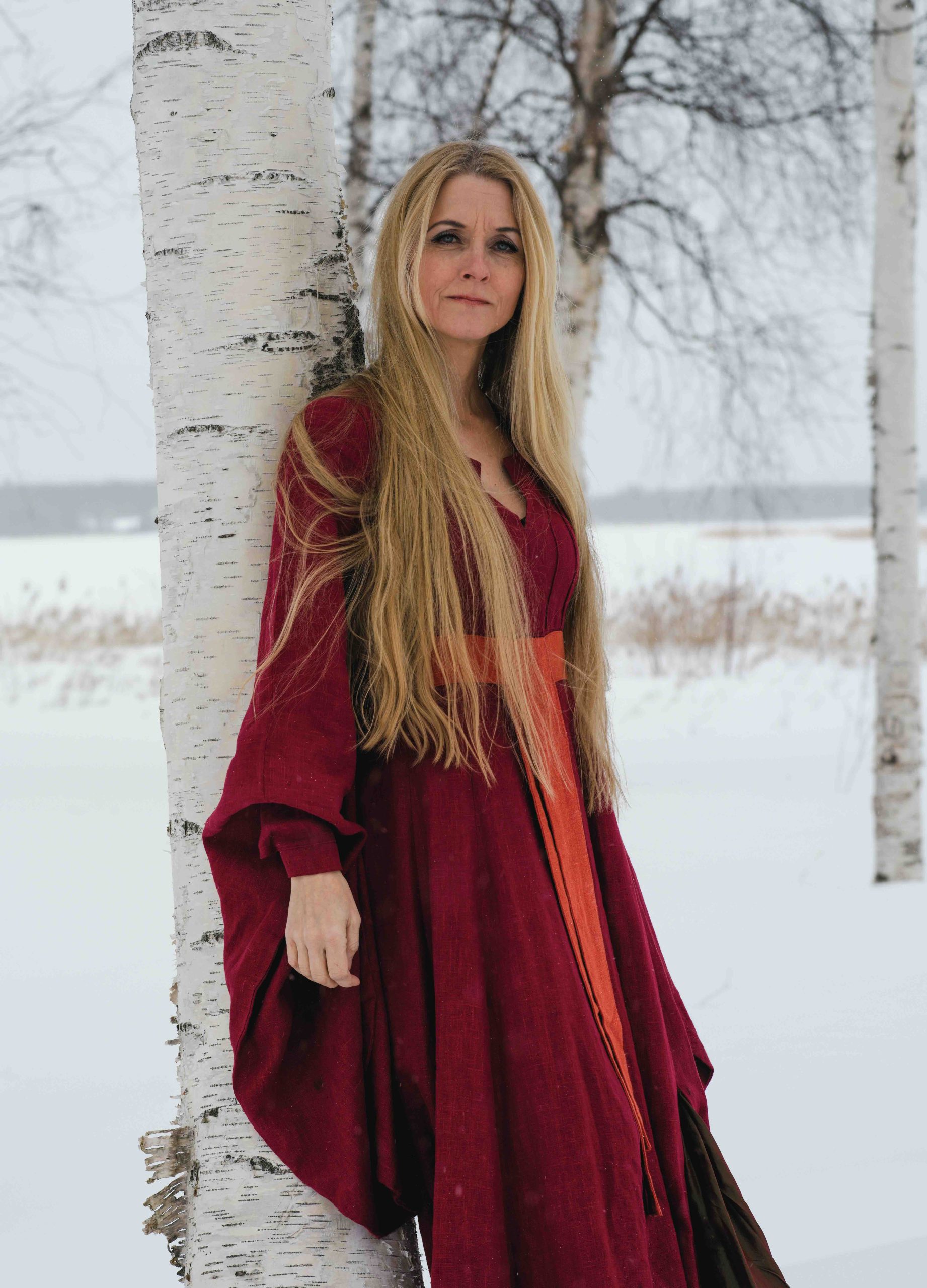 En kvinna i röd långklänning står lutad mot en björk i ett snöigt landskap. Hon har mycket långt, blont hår, och ler snett mot kameran.
