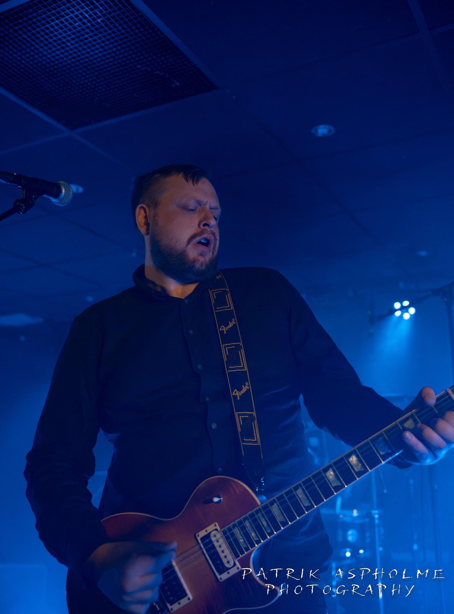 En kortklippt man i mörk skjorta spelar på gitarr med vidöppen mun.