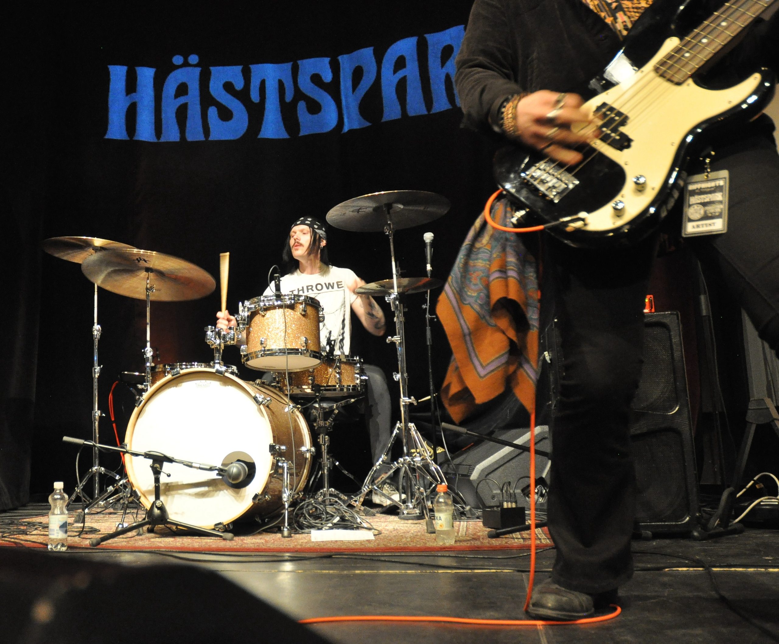 Hästsparks trummis i bakgrunden som spelar med blicken ner på trumsetet. Han har bandana och vit t-shirt och svart hår.