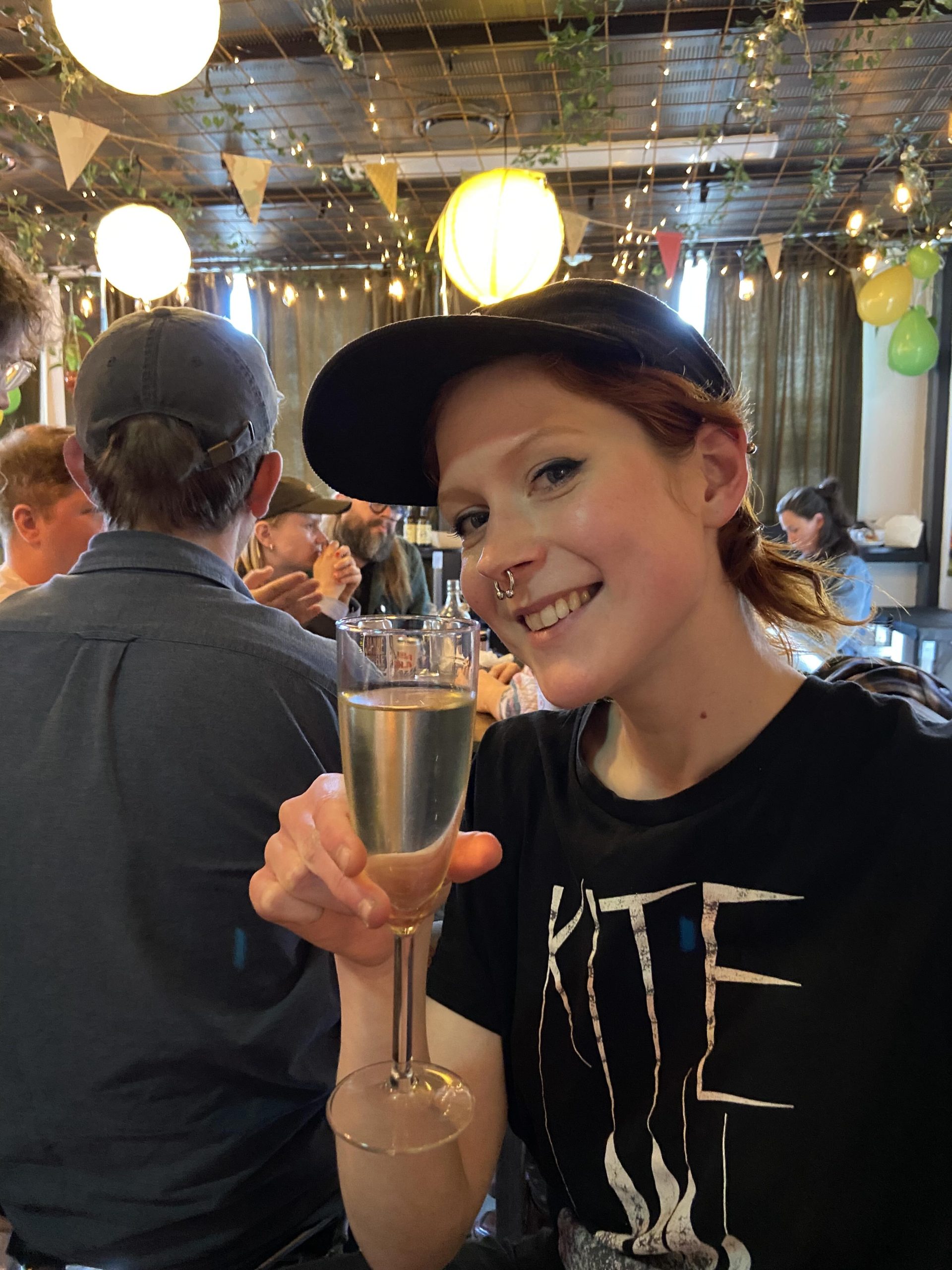Kvinna med keps ler mot kameran med ett glas cava i handen.