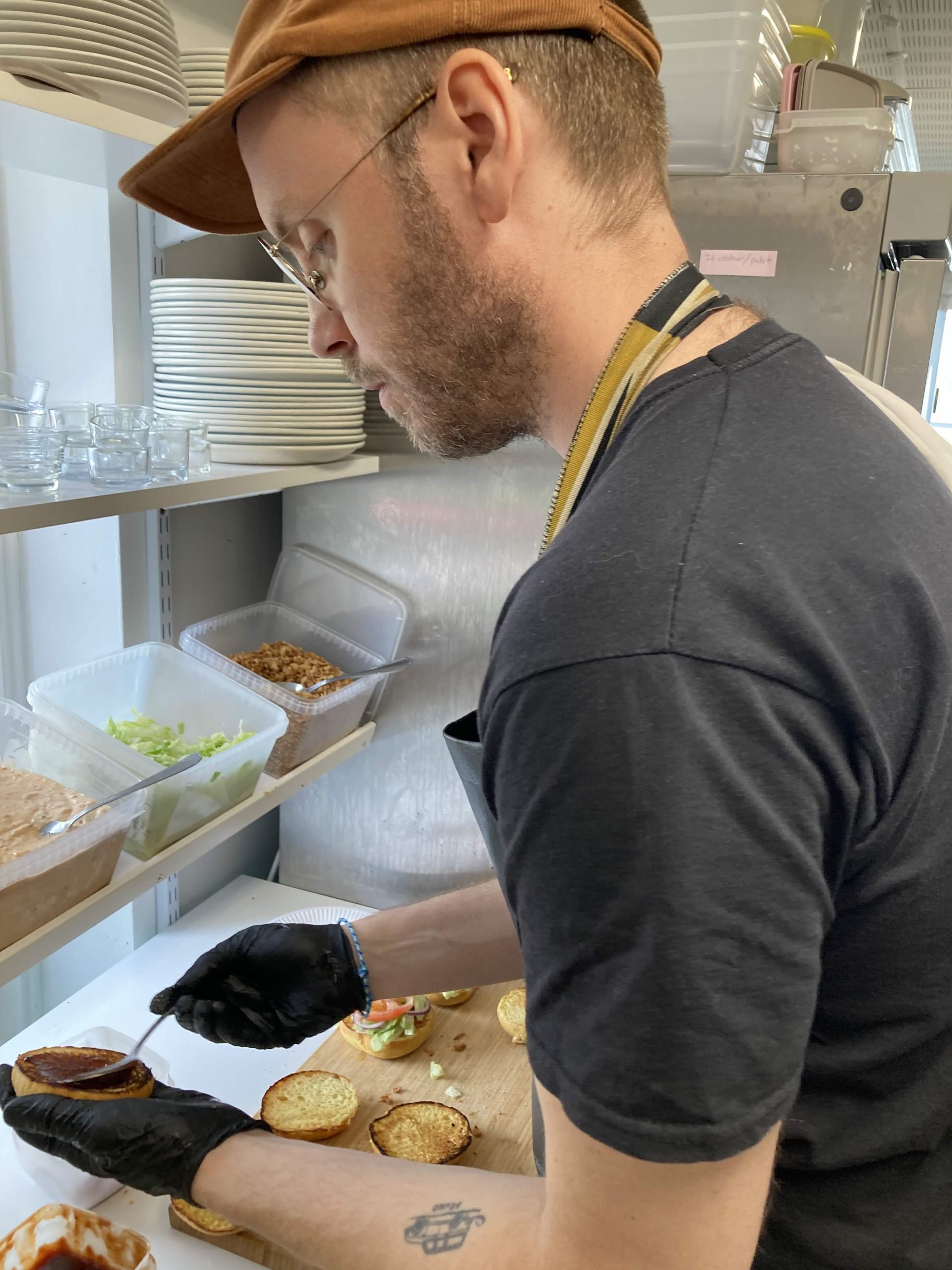En man med keps och glasögon och förkläde står och breder sås på burgarbröd. Framför honom står lådor med sallad, dressing och rostad lök.
