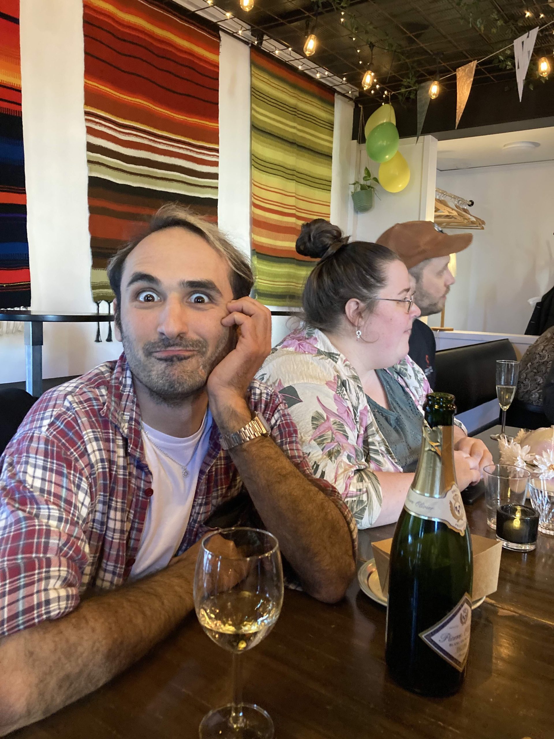 En man sitter vid ett bord med en cavaflaska och glas gör en grimas mot kameran.
