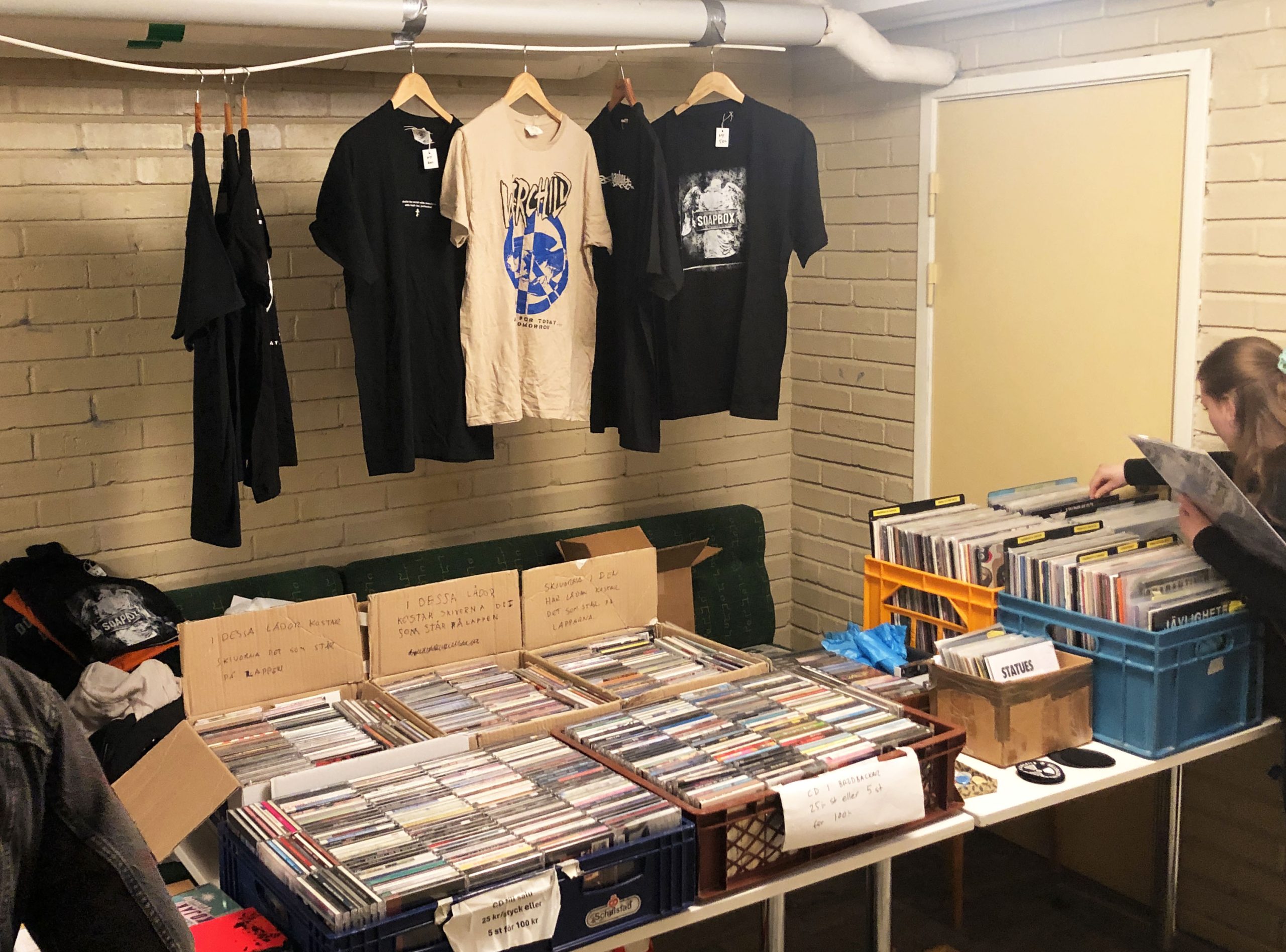 Ett långt bord med kasetter, CD-skivor och fyra backar med vinylskivor. En ung tjej står och bläddrar bland vinylerna. Det hänger också några t-shirts med olika bandtryck ovanför bordet.
