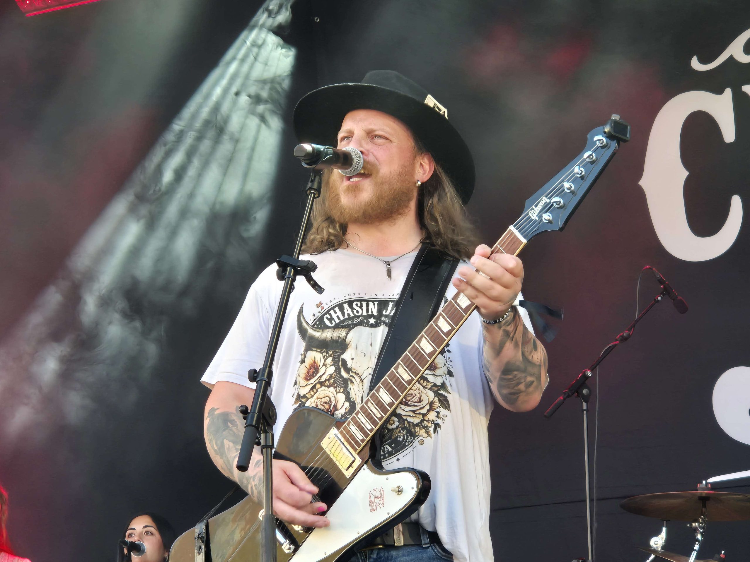 En kille med svart elgitarr, svart cowboyhatt och långt ljust hår. Han sjunger i mikrofonen och spelar sin gitarr vänd utåt mot publiken.