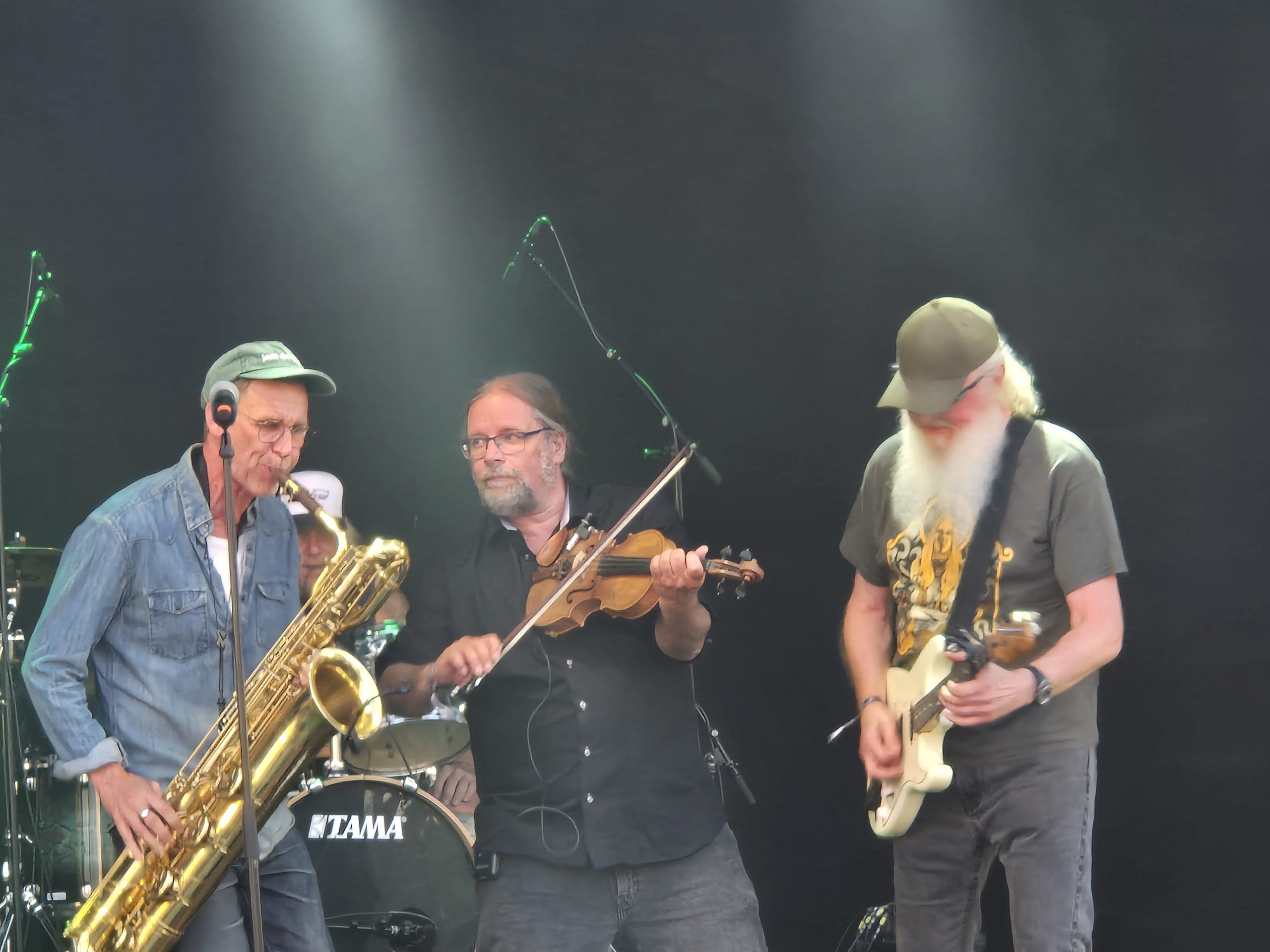 Tre gubbar på scen som spelar barytonsaxofon, fiol och elgitarr.