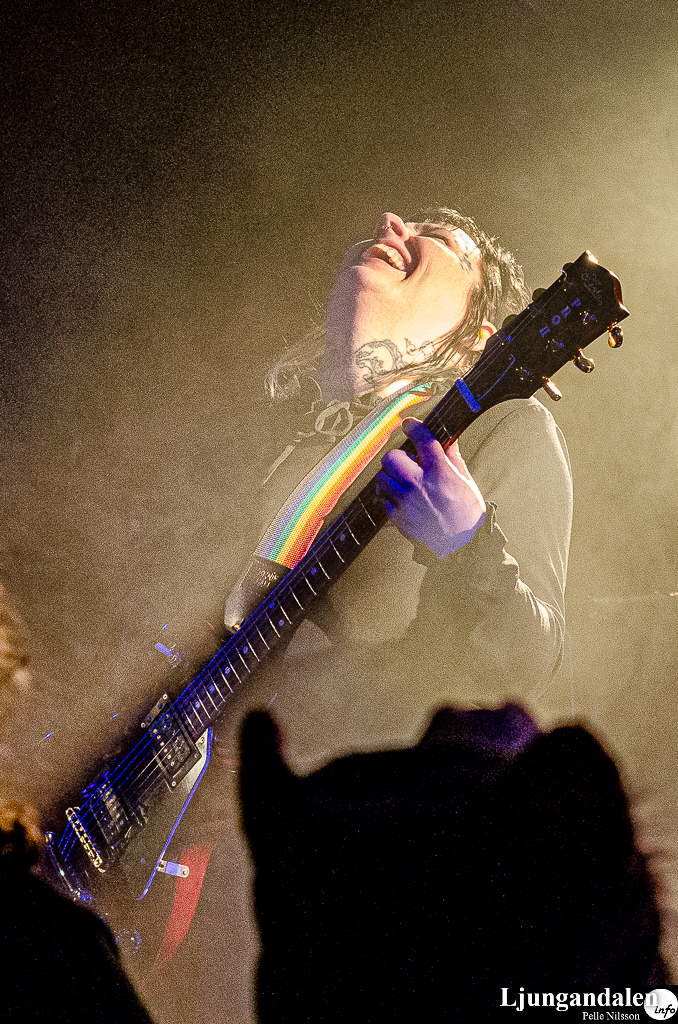 En kvinna med dreadlocks spelar gitarr och skrattar i dimmigt, blekt scenljus. Gitarrens axelband är priderandigt.