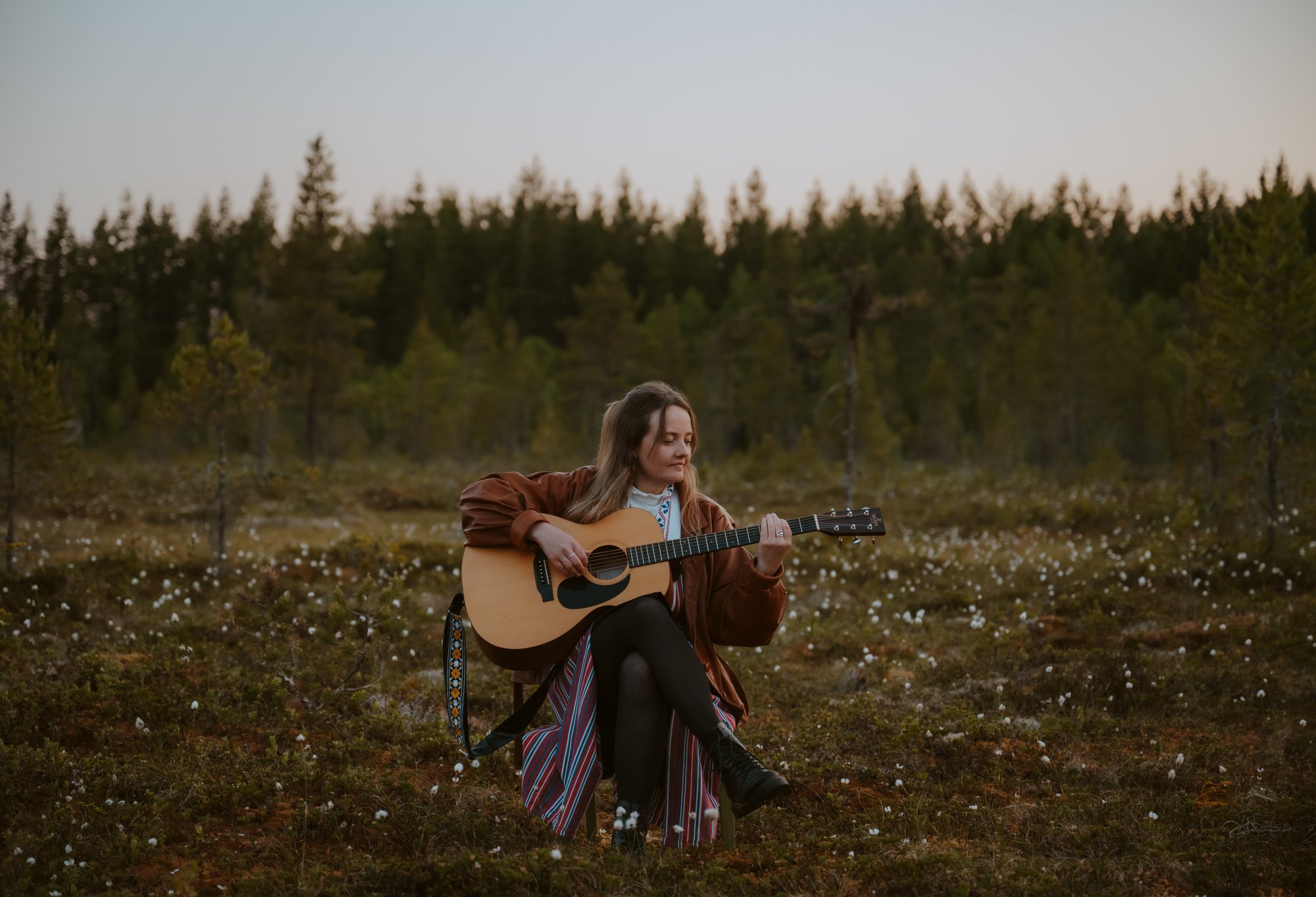 Artisten Ellen Sundberg sitter vid en myr i folkdräktsliknande utstyrsel och spelar på en akustisk gitarr.