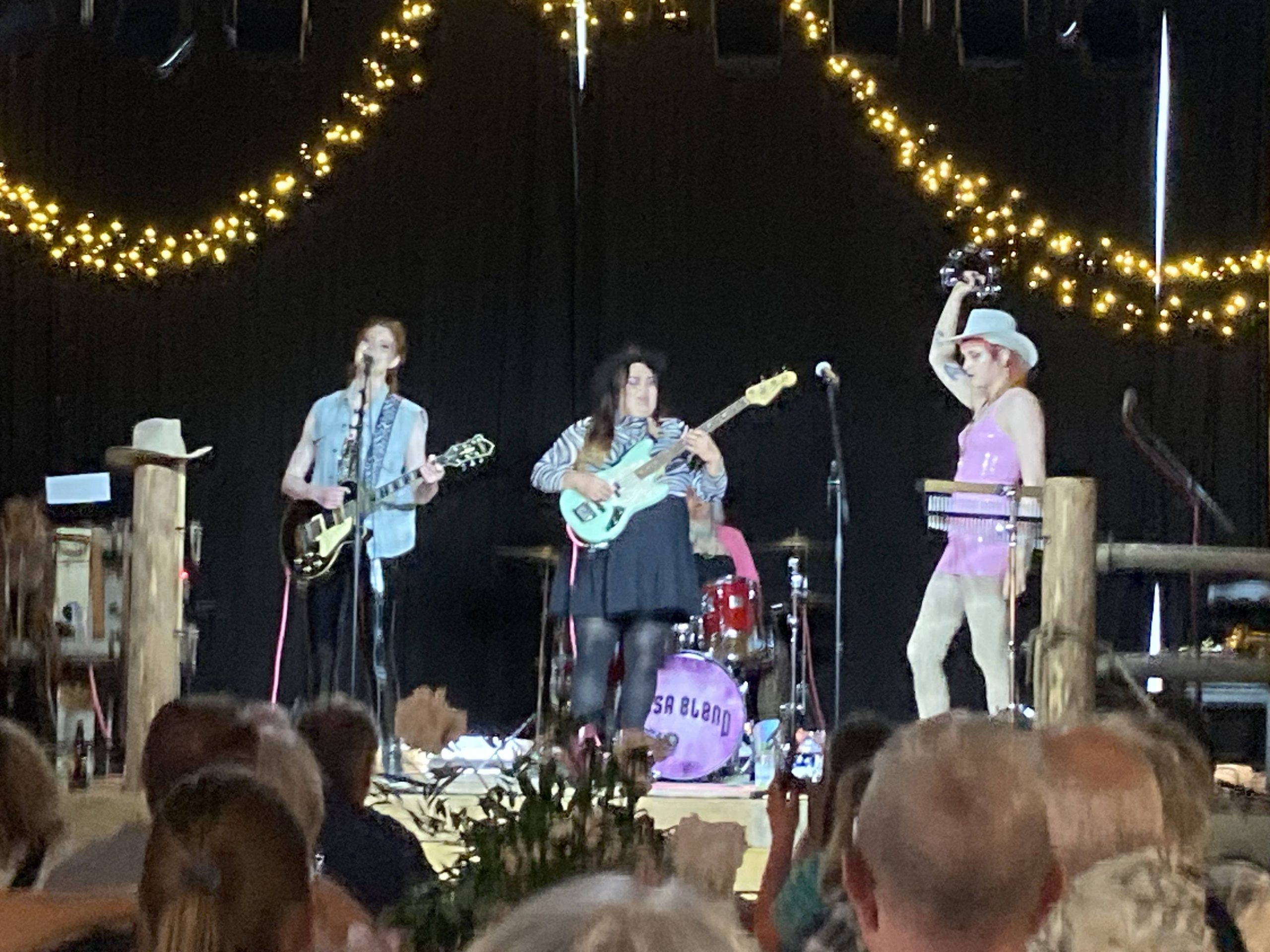 Fyra tjejer på scenen med elgitarr, elbas, sångmick och tamburin. Längst bak skymtas trummisen. Ena tjejen har kort, rosa lackklänning och en vit cowboyhatt.