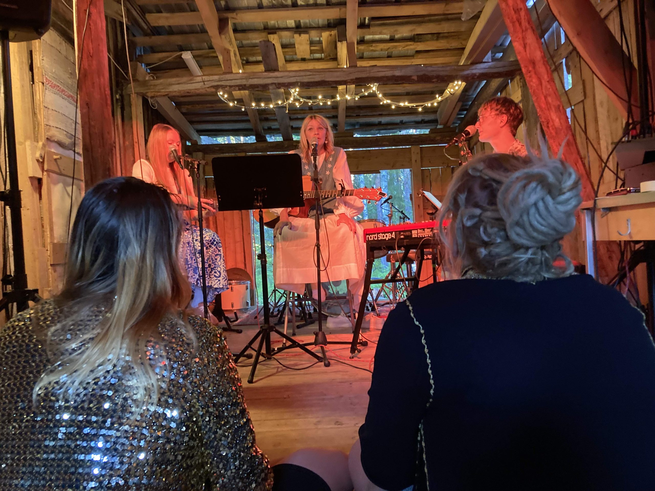 En konsert i en lada. Två personer sitter på golvet framför de tre musikerna som spelar musik med sång, keyboard och två akustiksa gitarrer, två kvinnor och en man.