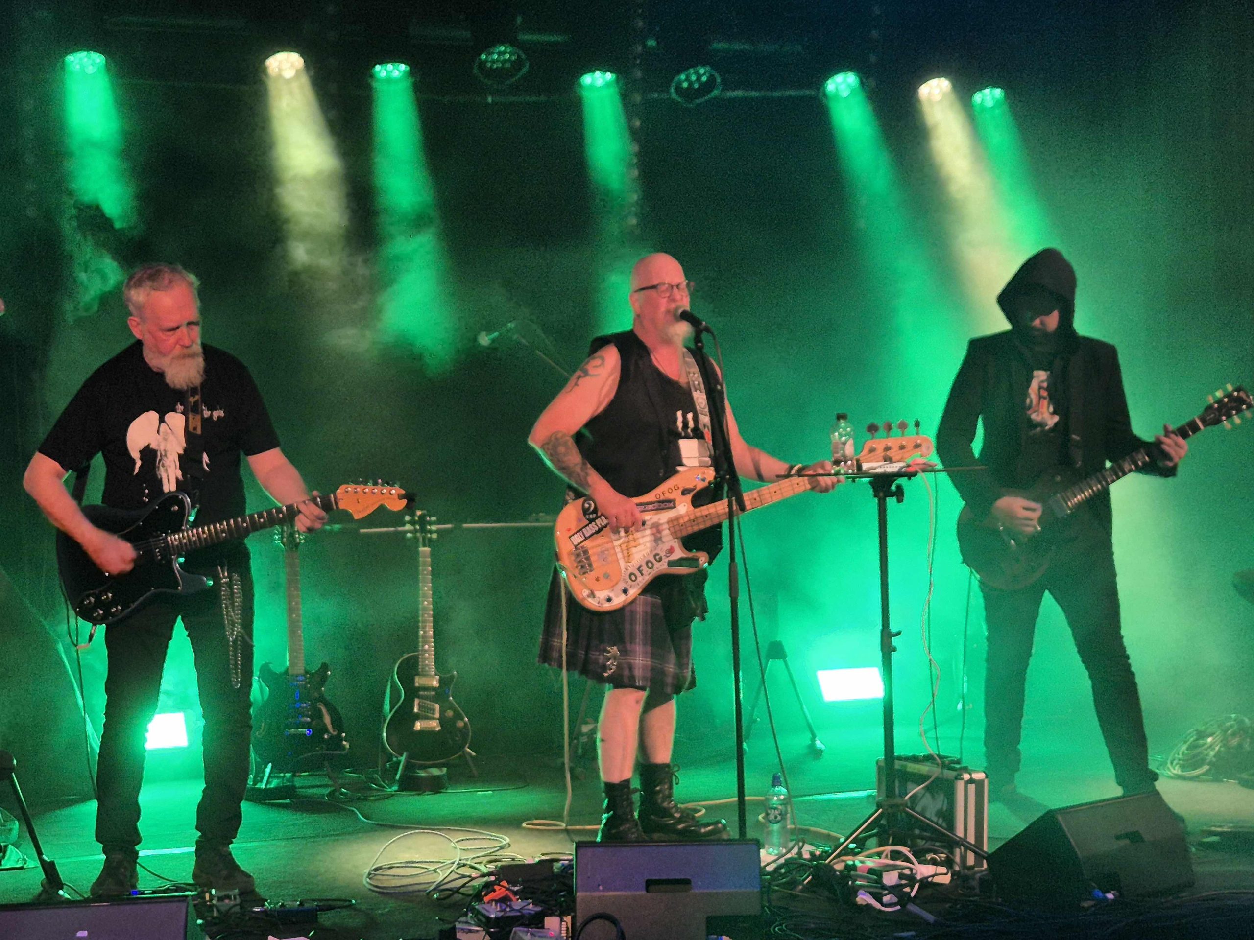 Bandet O.F.O.G. Tre män i 60-årsåldern står på scen och spelar elgitarr och elbas i grönt scenljus med mycket rök. Mannen till höger har svart kavaj och luva uppdragen över huvudet. Mannen i mitten har en väst och kilt och vitt skägg, och mannen längst ut till vänster har mörk t-shirt och jeans och grått skägg.