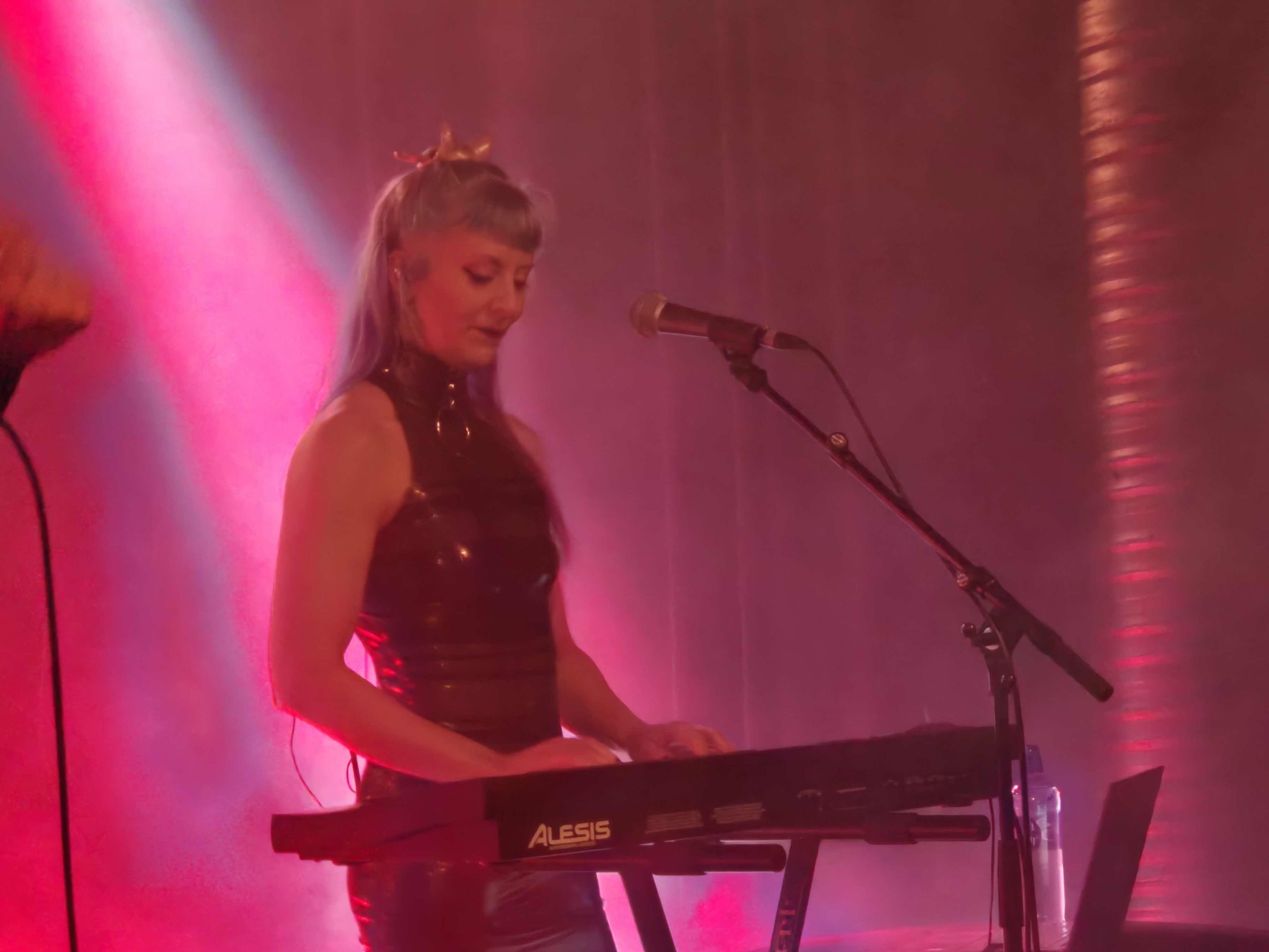 En kvinna som spelar keyboard. Hon ler ner på tangenterna medan hon spelar. Hon har en svart latexklänning på sig och blått hår. Scenbelysningen är starkt rosa.