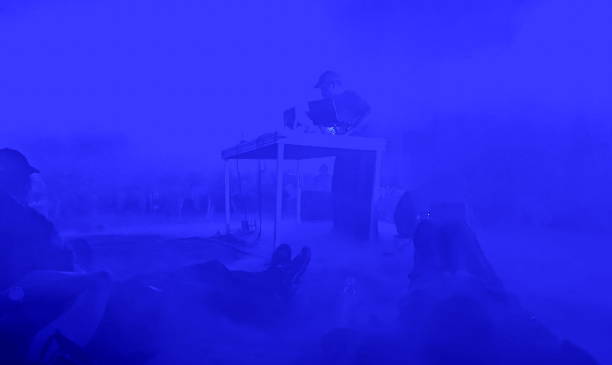 Blått scenljus och rökmaskin skapar en dimmig effekt. En DJ står vid ett DJ-bord, runt honom kan man urskilja några personer som ligger ner i röken.