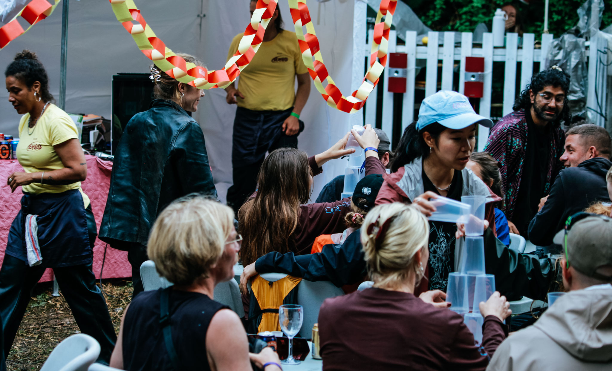 Festivalbesökare som gör olika grejer. En ung kvinna i ljusblå keps och svart långt hår tar en plastmugg från bordet. Hon och de andra runt bordet bygger ett torn av muggarna. Bakom dem är det annat bord där deltagarna bygger torn.
