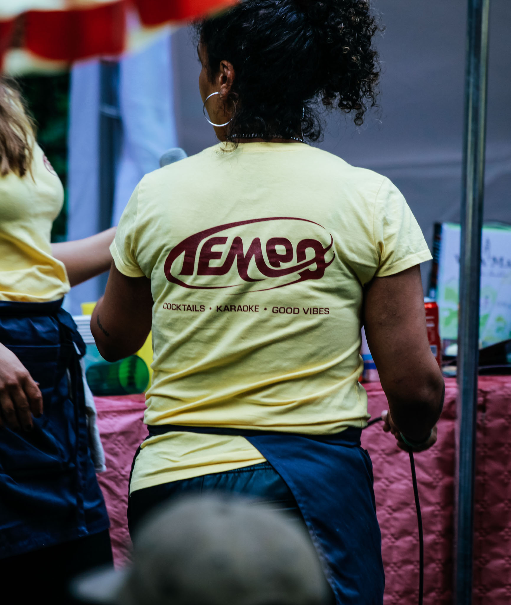 En mörkhyad kvinna med stora silverringar i öronen står med ryggen vänd mot kameran. Hon har ett förkläde och en gul t-shirt med texten "TEMEO - cocktails, karaoke, good vibes" på ryggen.