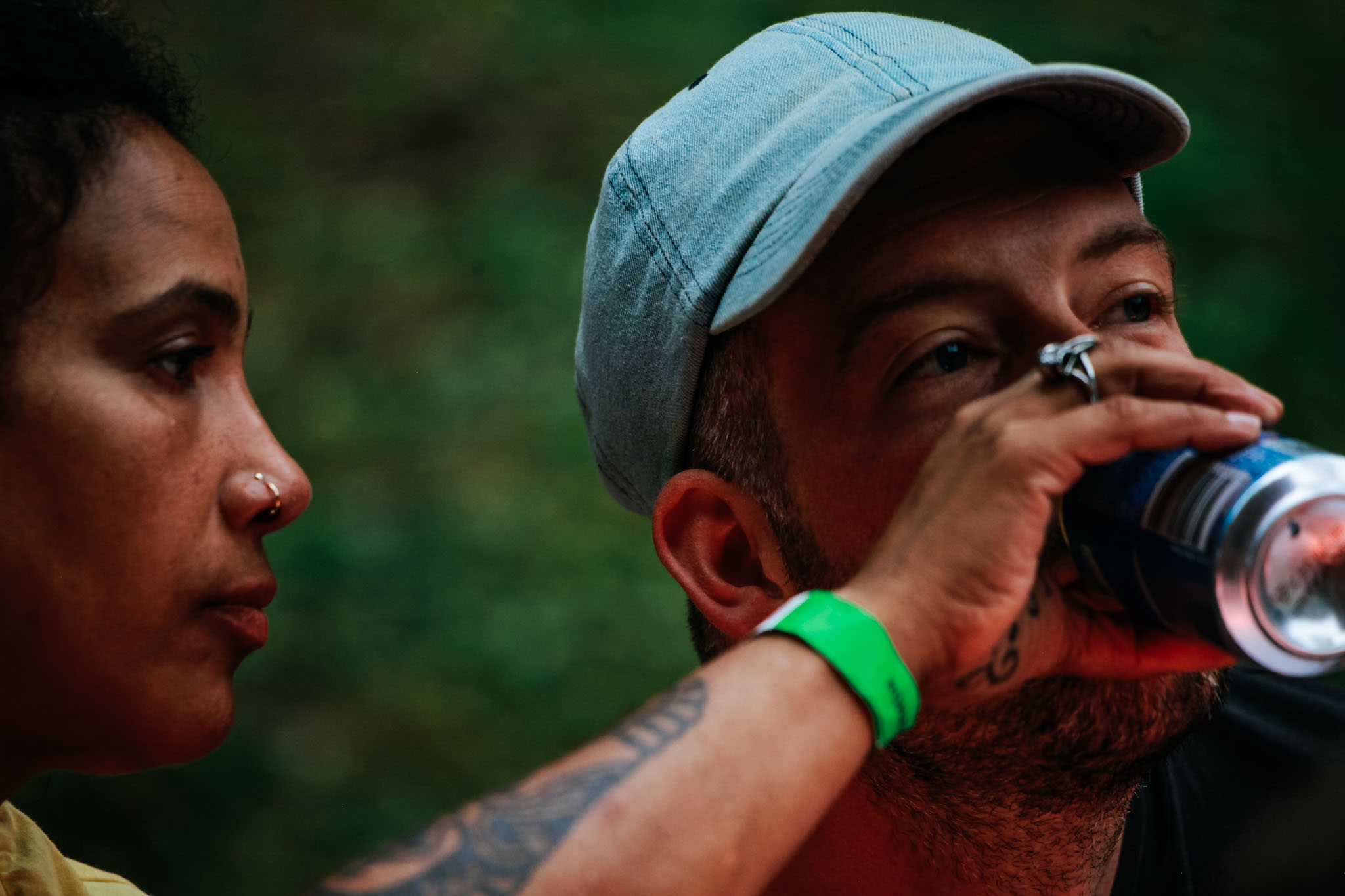 En kvinna med piercing i näsan häller en burk öl i en mans mun. Mannen är en ljusblå keps. Kvinnan har ett illgrönt festivalarmband runt handleden.