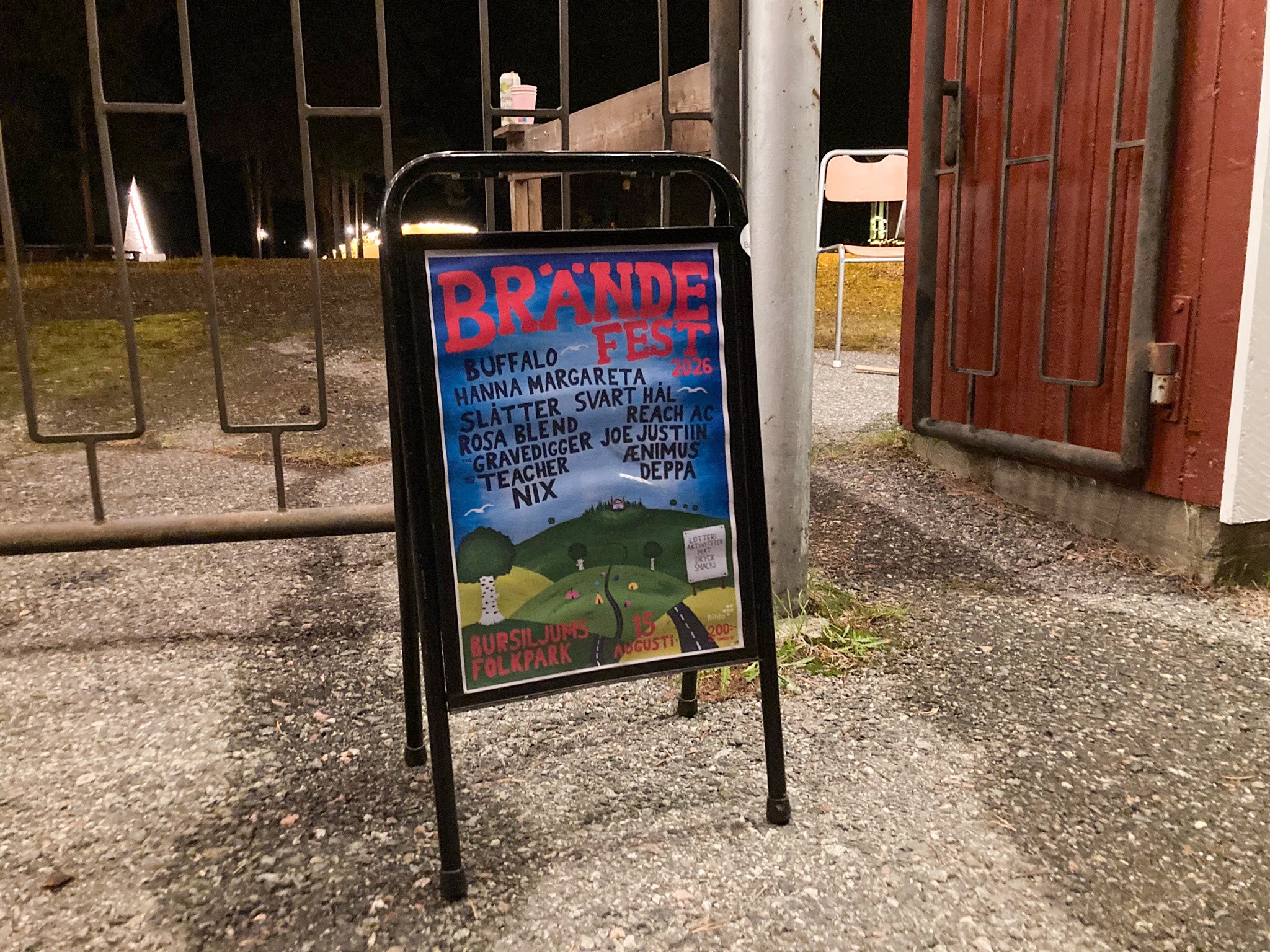En trottoarpratare med en affisch utanför en grind. På affischen står det BRÄNDEFEST 2026 i stora bokstäver och under alla bandnamn. I bakgrunden är en illustration av ett böljande landskap.