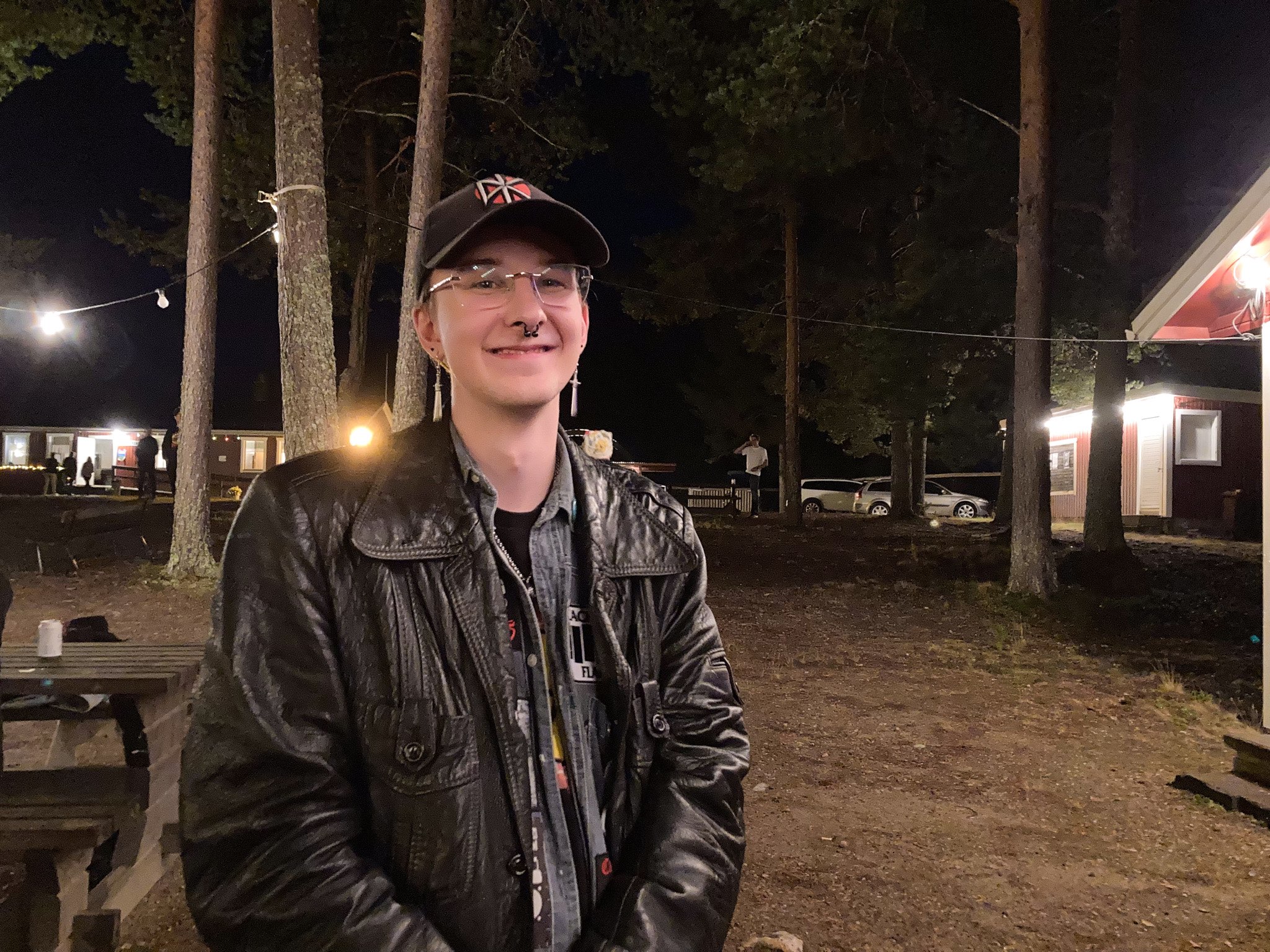 En ung kille med skinnjacka, glasögon, piercing i näsan och svart keps. Han ler in i kameran. Bakom honom syns folkparken, det är mörkt ute.