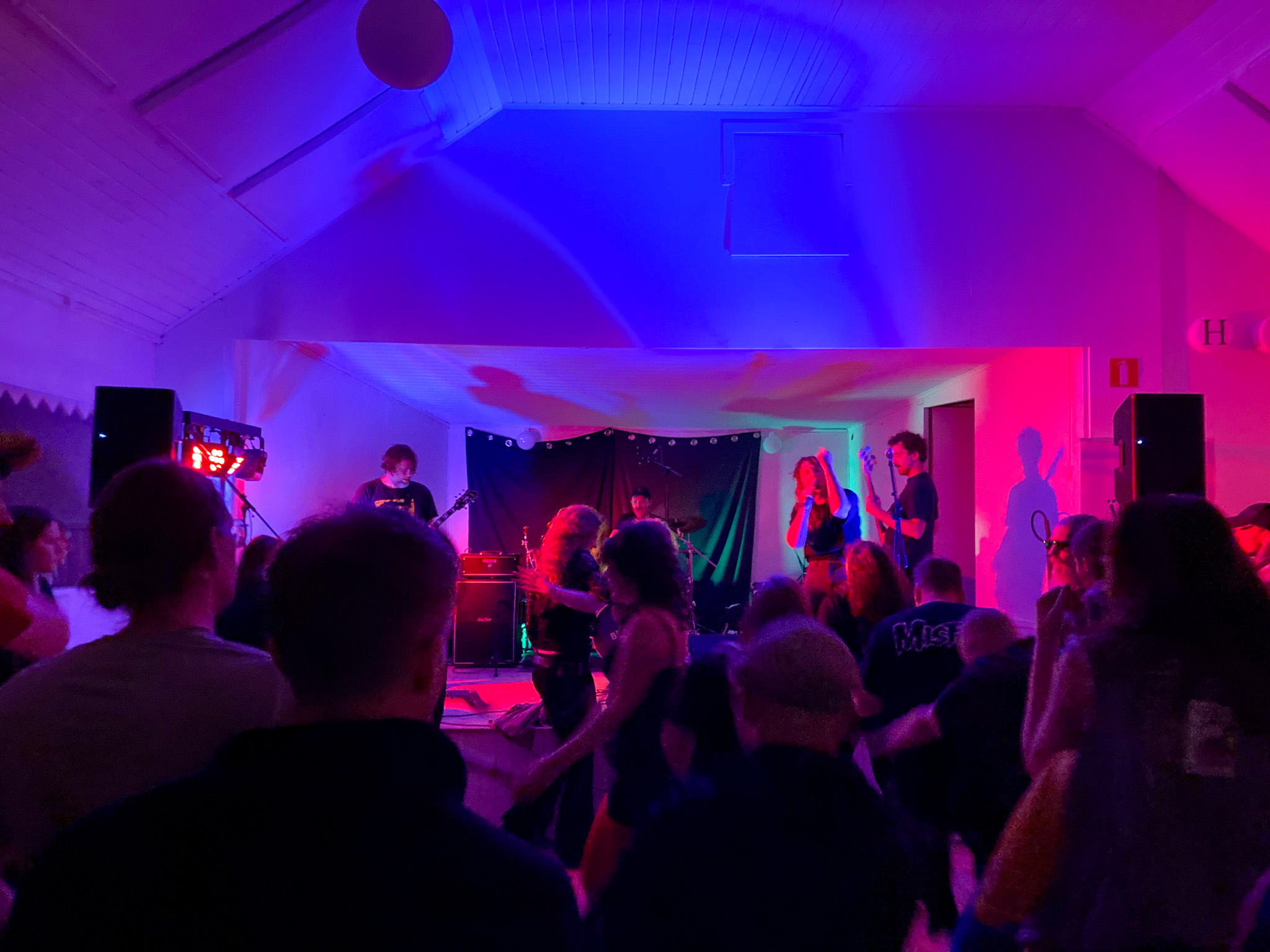 Inuti en danslada. Taket och väggarna är vitmålade. På scen står ett band och framför dansar publik. Scenljuset är starkt blått och rosa.