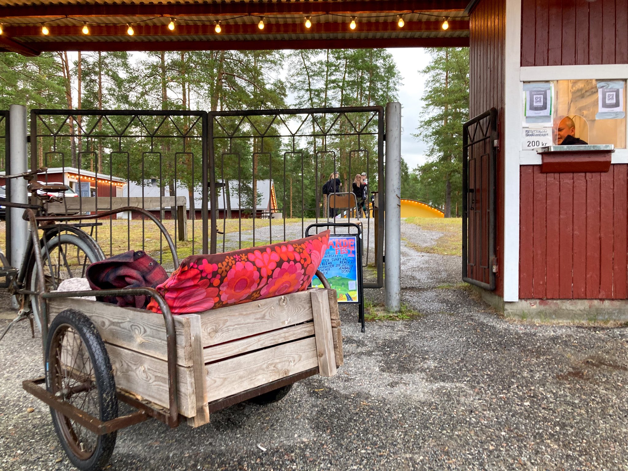 Utanför folkparkens entré. En cykel med vagn är parkerad utanför grinden. I vagnen ligger en färgglad kudde och en flanellskjorta.