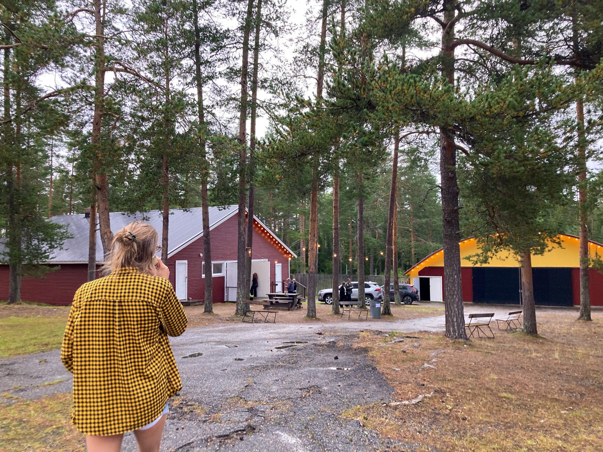 En person med långt, ljust hår uppsatt i en slarvig knut och med korta shorts och gul- och svartrutig skjorta går på området med ryggen vänd mot kameran. På området syns höga tallar och två rödmålade byggnaden pyntade med ljusslingor.