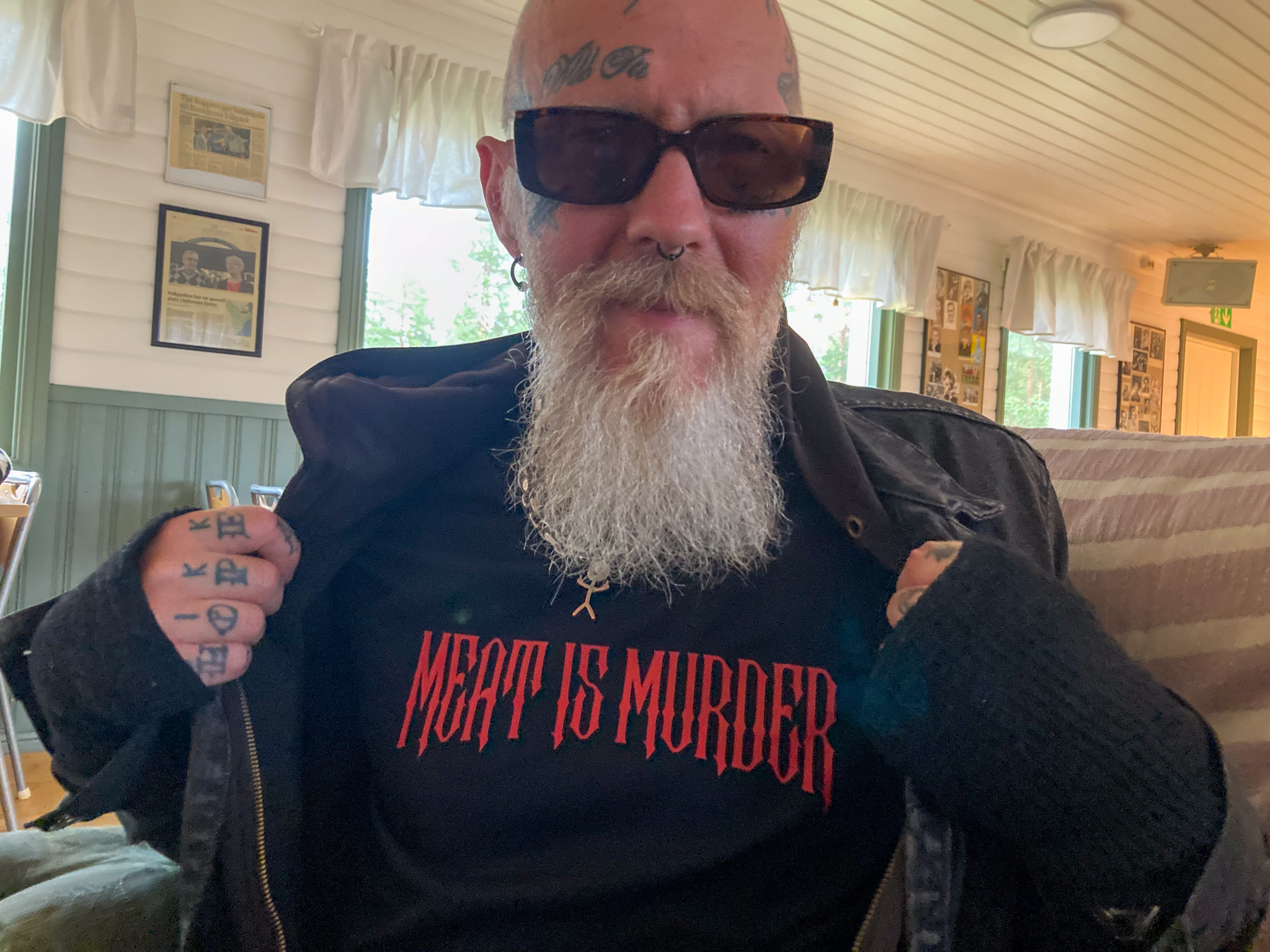 En man med tatuteringar i ansiktet, piercing i näsan, långt vitt skägg och solglasögon ler in i kameran. Han drar undan sin jacka för att visa sin t-shirt, som det står "Meat is murder" på.