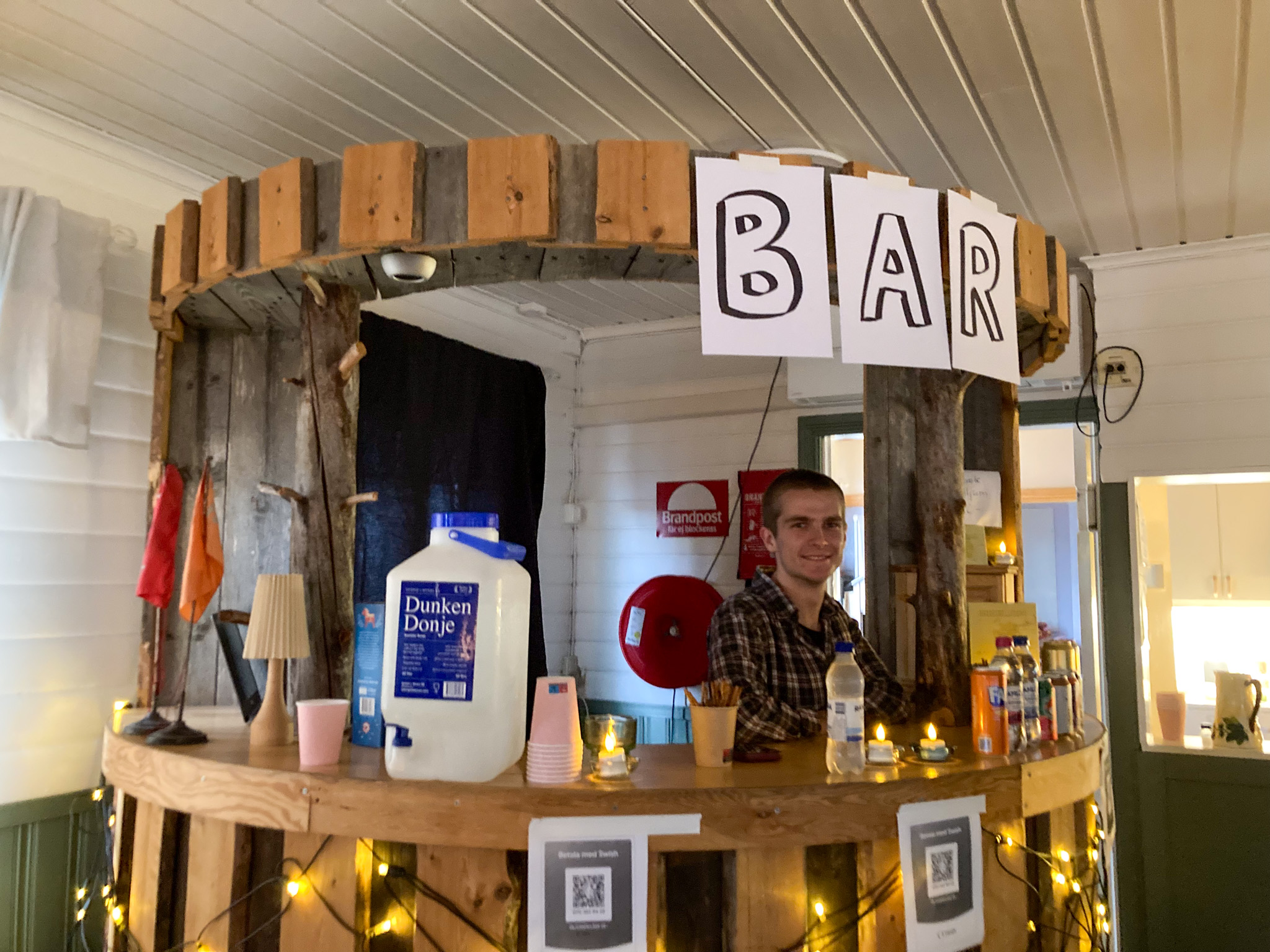 En bar snickrad i en halvcirkelform. I baren står en ung, snaggad man med rutig skjorta och ler mot kameran. På baren står en dunk med vatten, pappmuggar, en lokaflaska, läsk, öl och cider. Längst upp på baren sitter tre A4-papper upptejpade, som tillsammans bildar ordet "Bar".