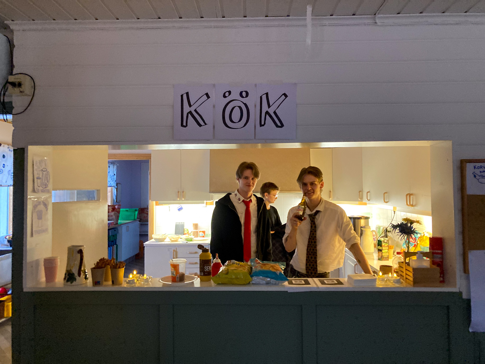 Tre unga personer står i en lucka. Längst upp sitter tre A4-papper upptejpade, som tillsammans bildar ordet "Kök".