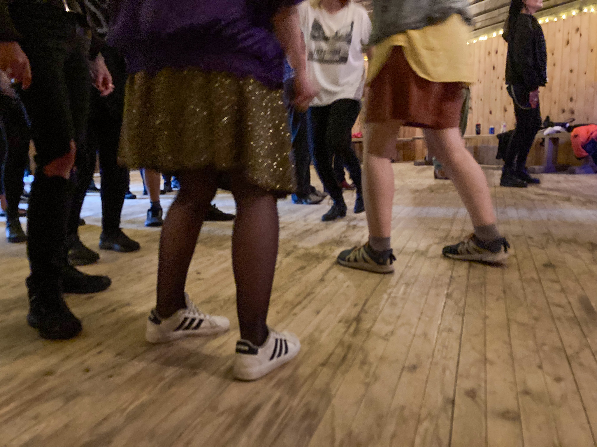 Fötter som rör sig på ett trägolv. I förgrunden är ett par vita gympaskor, benen är klädda i svarta nylonstrumpbyxor och en guldglittrande kjol.