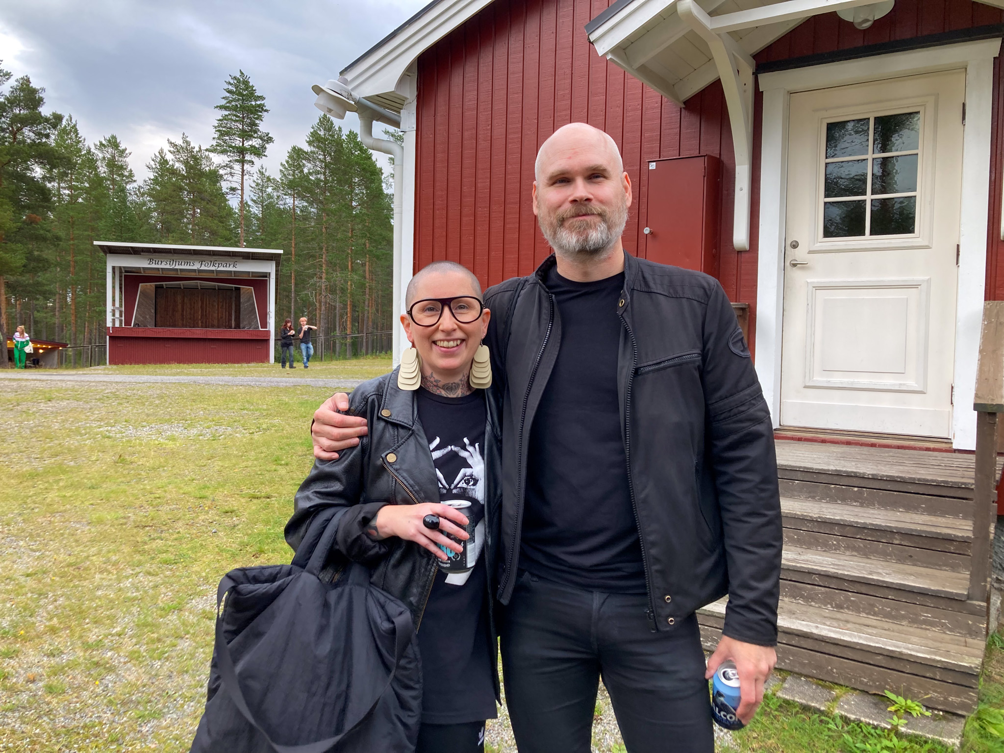 Två personer står framför en av byggnaderna på området och ler mot kameran. Till vänster en kvinna med stora glasögon, skinnjacka, stora örhängen och rakat huvud. Till höger står en lång man med svarta kläder, grått skägg och rakat huvud. Han håller armen om henne. Båda håller en varsin ölburk.
