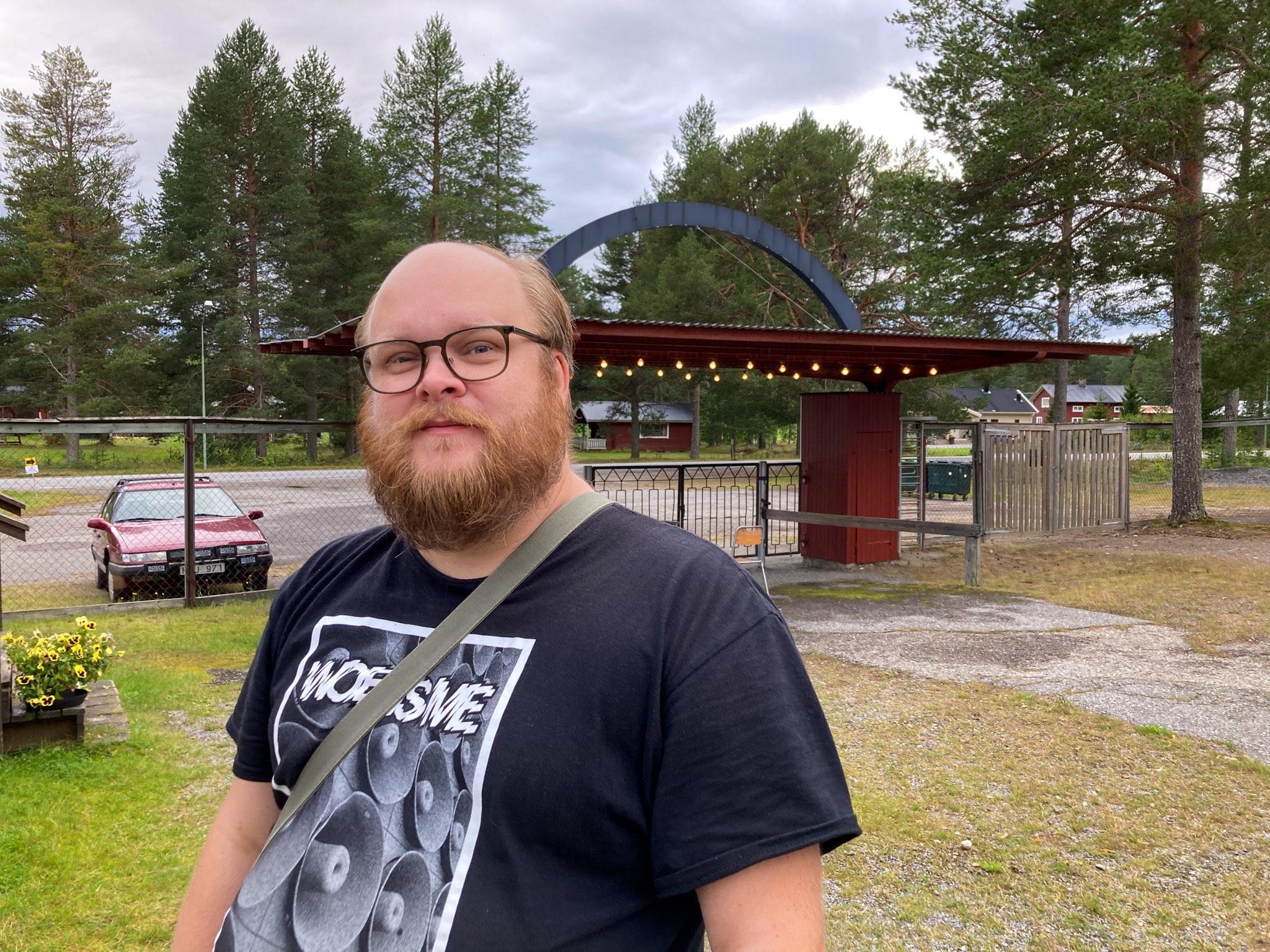 En man med ljust skägg, glasögon och bandtröja ser in i kameran. Bakom honom syns folkparkens entré.