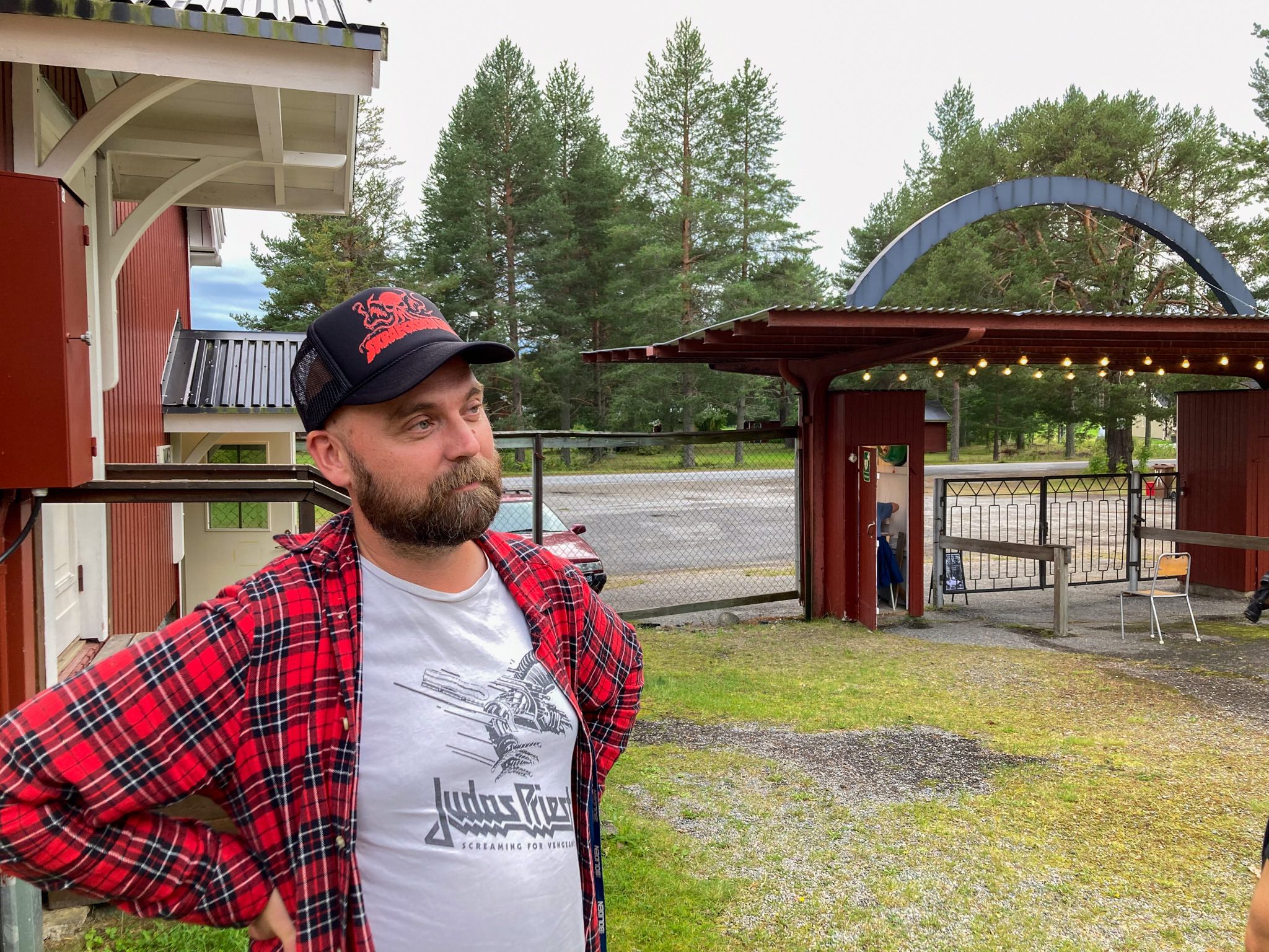 En man med mörkt skägg, keps, rödrutig flanellskjorta och en Judas Priest-tröja. Han står med armarna i sidorna och ser bortåt. Bakom honom syns folkparkens entré.