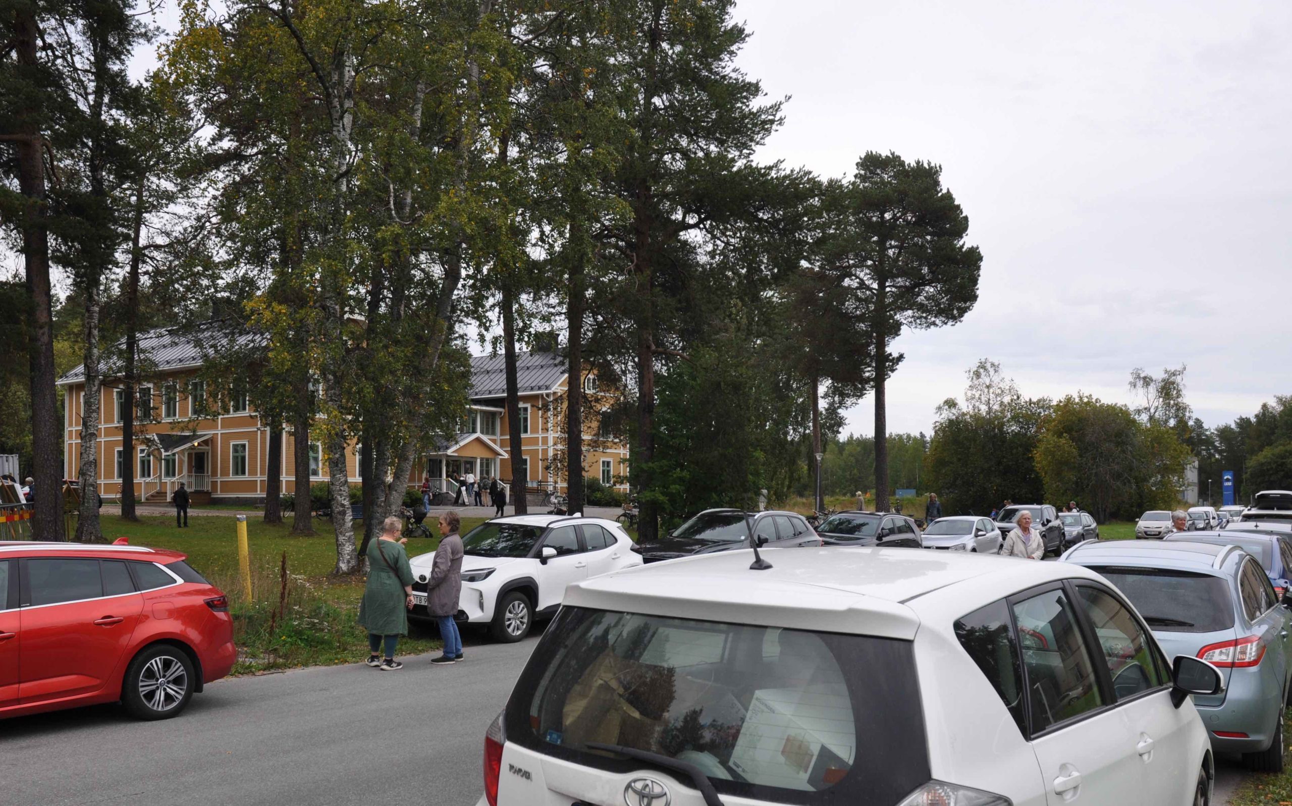 Långa rader av parkerade bilar och gamla människor som står och pratar med varandra. I bakgrunden syns Blackis bakom några träd.