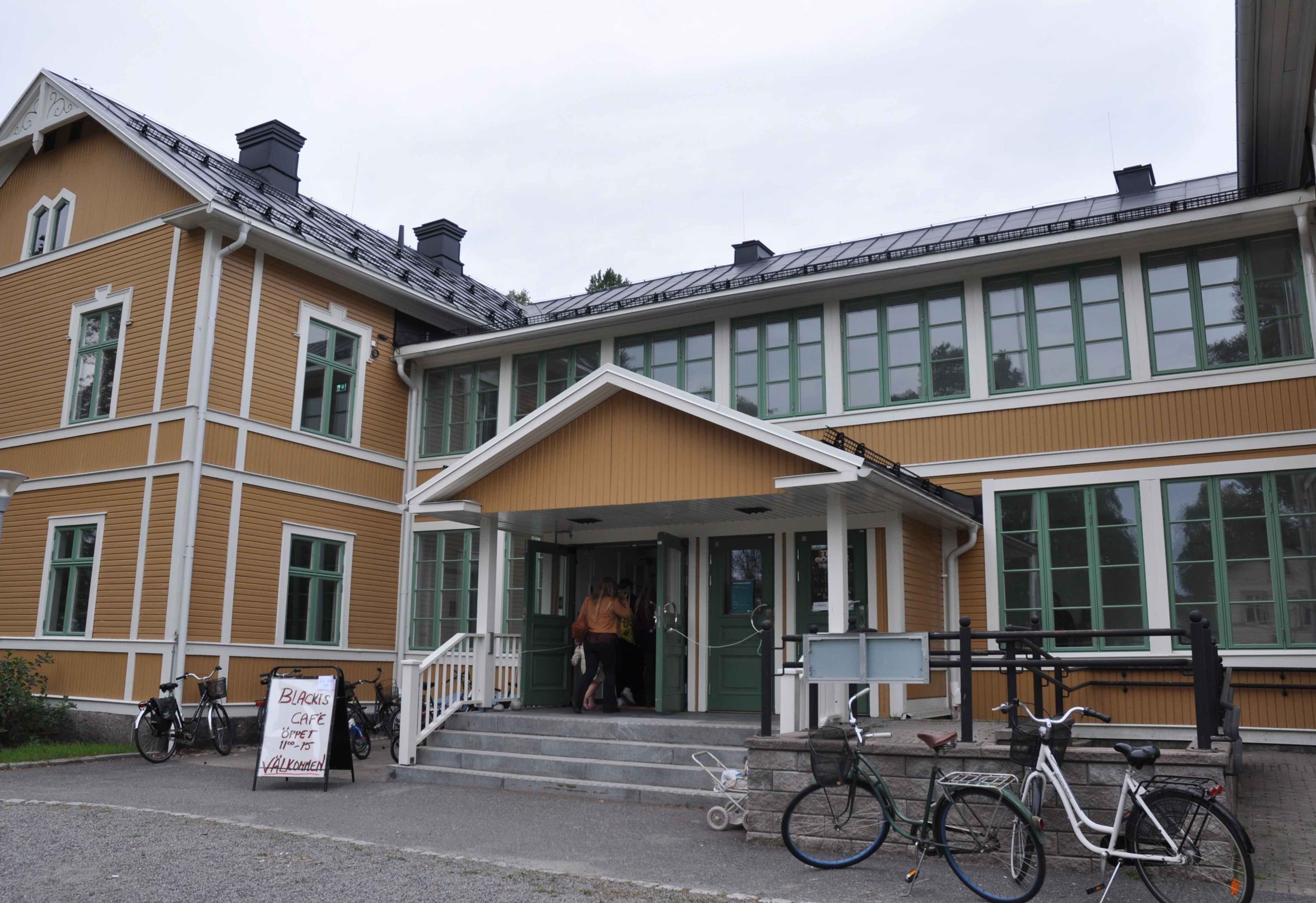 Blackis entré. Två kvinnor är påväg in i byggnaden. Utanför står en skylt med texten "Blackis café öppet 11:00 till 15:00