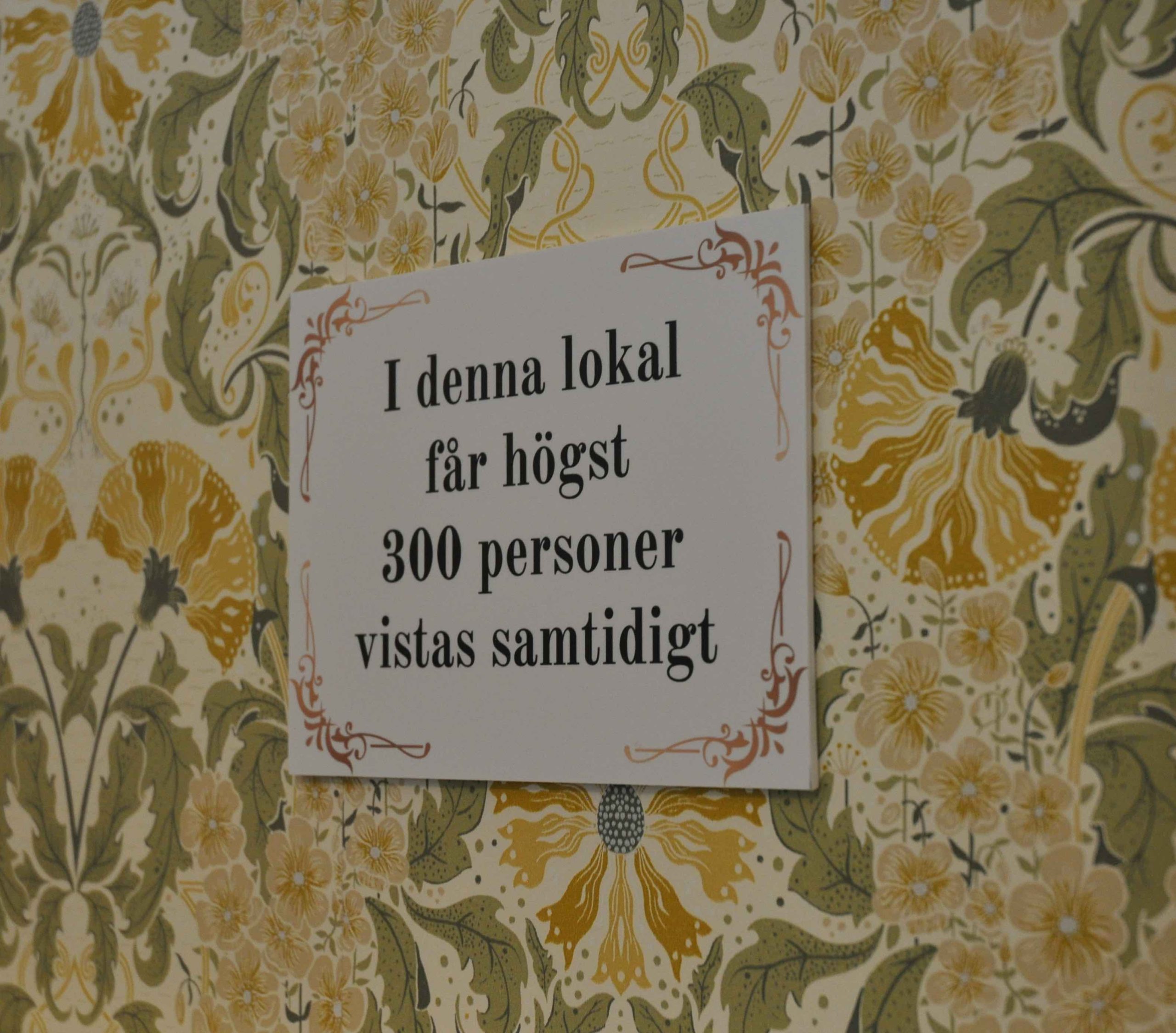 En skylt uppsatt på en gulblommig retrotapet. På skylten står det "I denna lokal får högst 300 personer vistas samtidigt".
