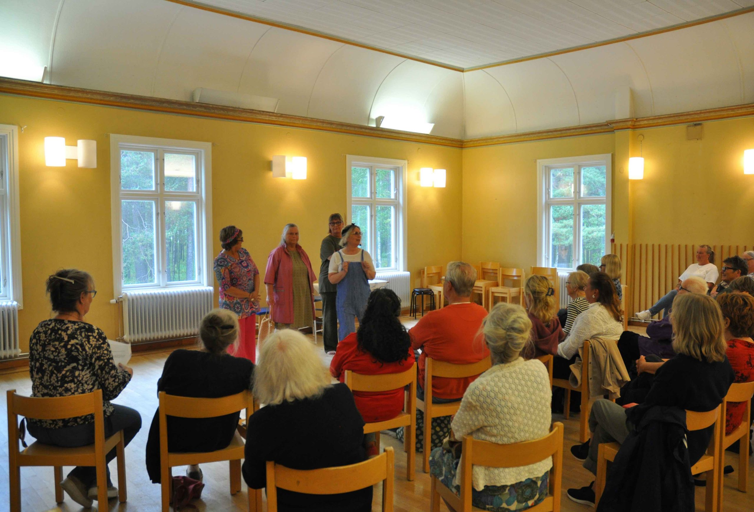 Ett gäng äldre damer står framför en äldre sittande publik på ca 20 pers. Damerna verkar framföra teater utan rekvisita.