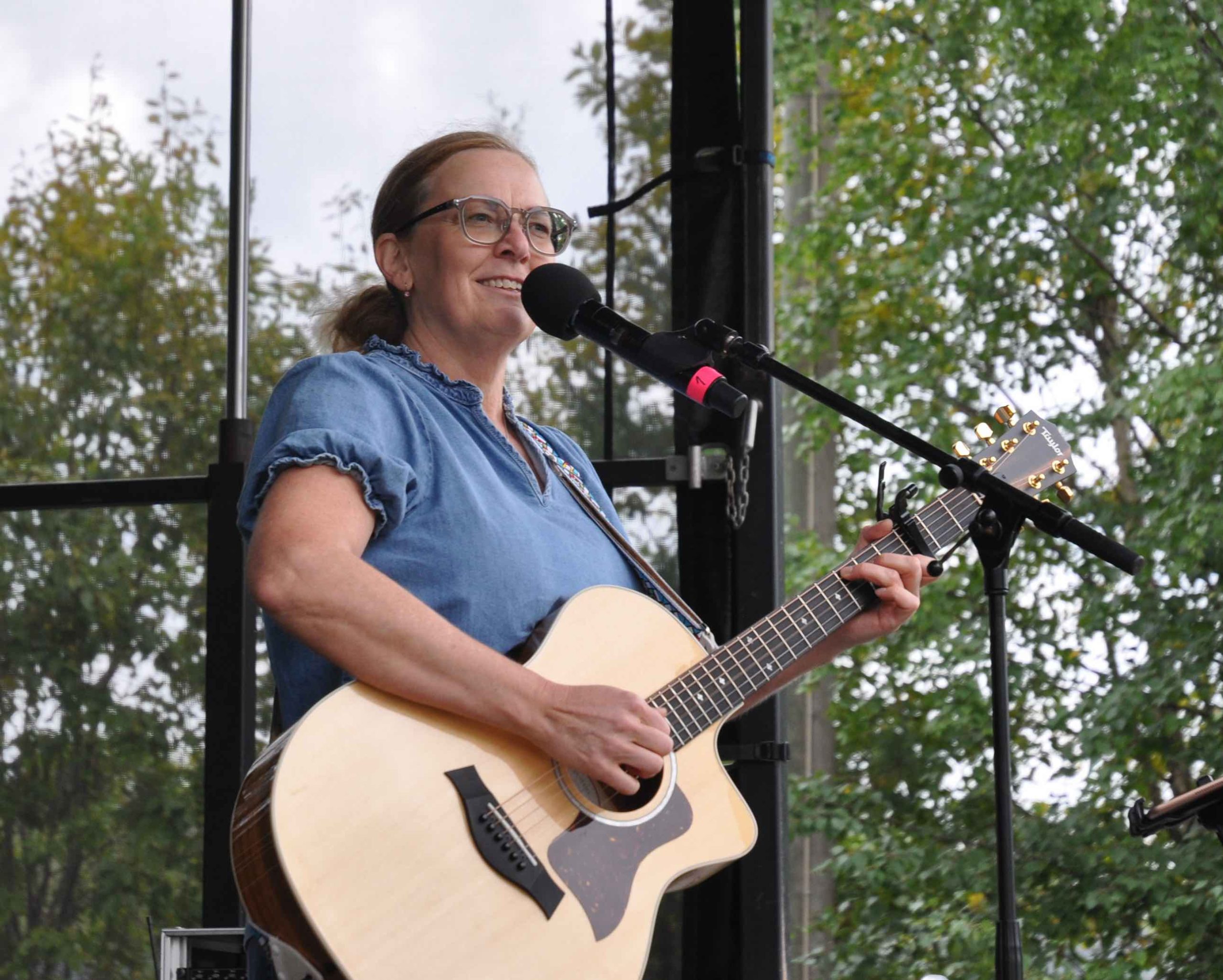 Artisten Anna Sellén står på scen med sin gitarr och ler mot publiken. Anna har mörka glasögon, en blå top och brunt hår i en lågt uppsatt hästsvans.